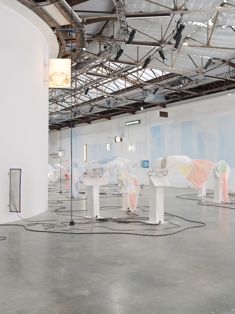 Wide installation view of white pedestal sculptures covered in pastel translucent fabric beneath an industrial skylit ceiling.