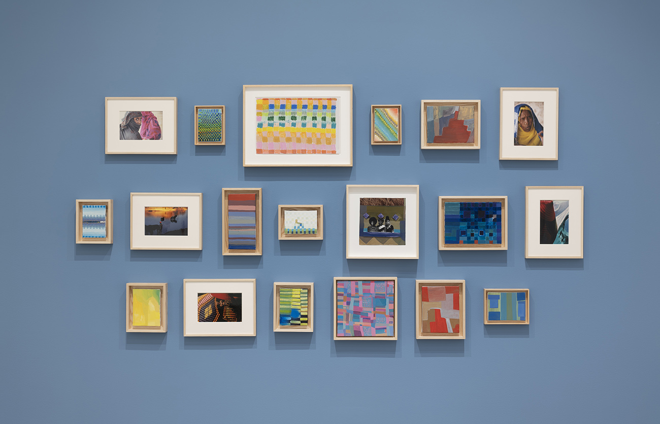Gallery wall featuring a grid of small colorful abstract paintings and portraits against a blue wall.