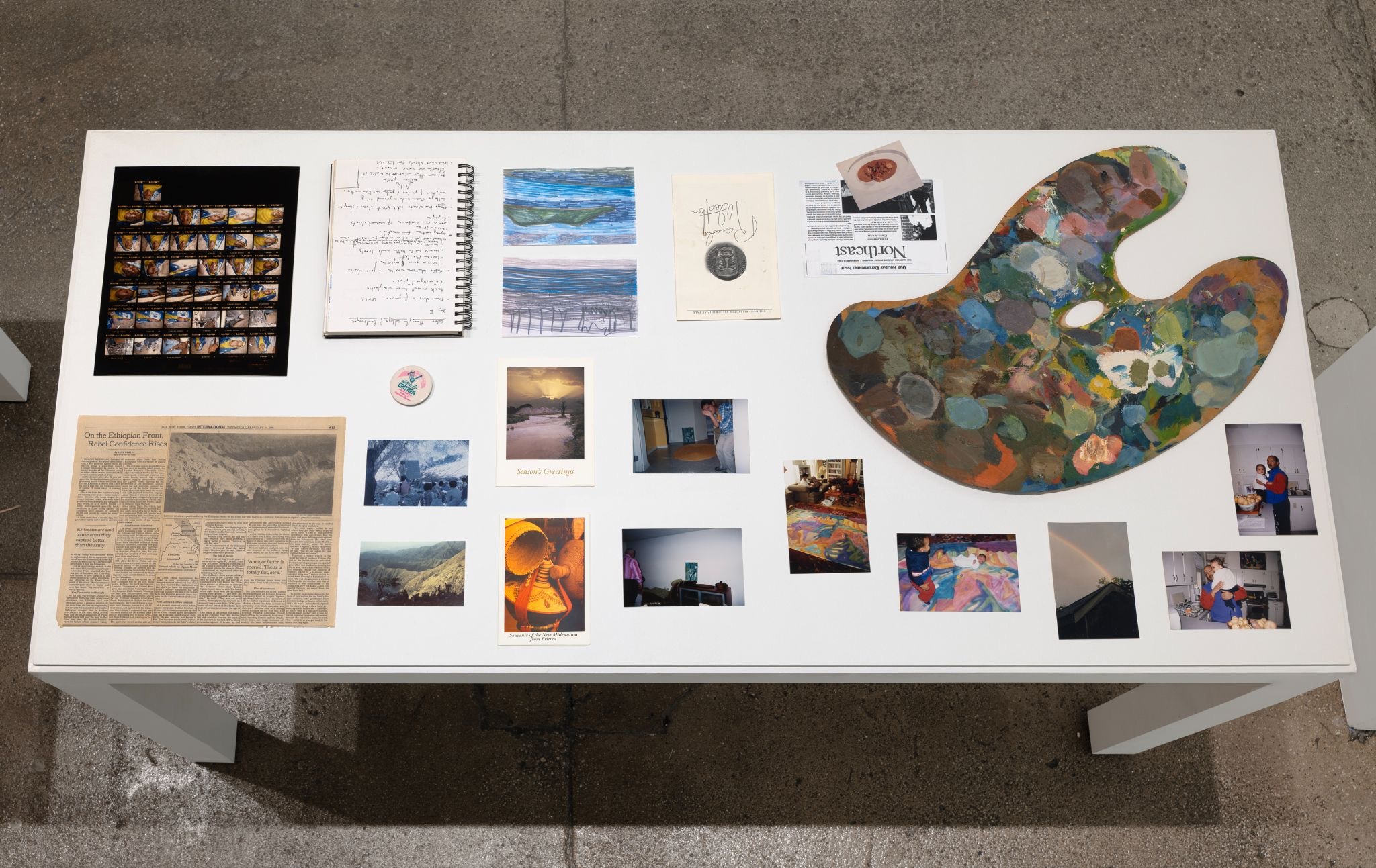 Exhibition table displaying photographs, sketches, printed materials, and a painter’s palette covered in multicolored paint.