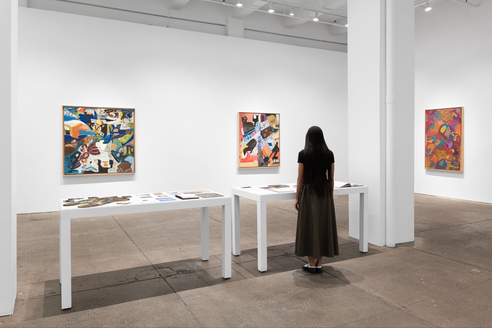 Visitor standing in a white gallery space viewing large colorful abstract paintings and display tables.
