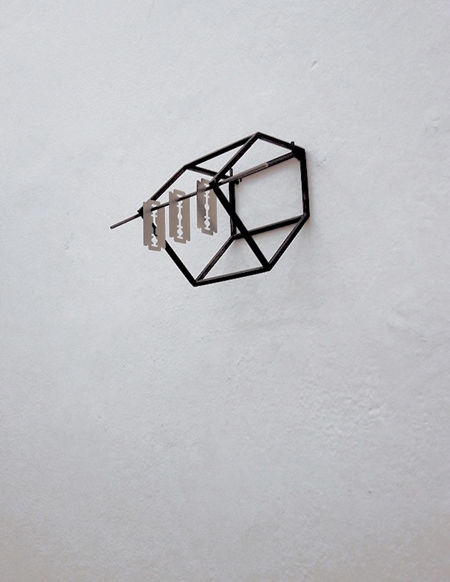 Three razor blades hang from a welded metal geometric shape mounted on a wall.l