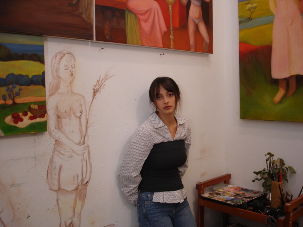 Female artist stands against a white wall with paintings hung around her.