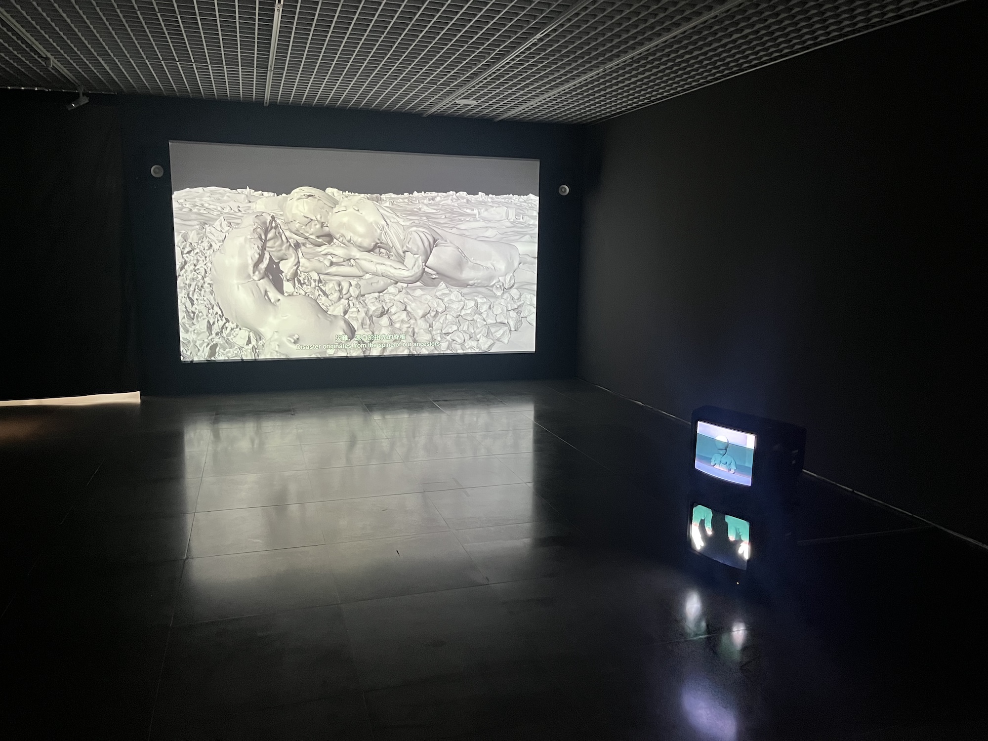Video installation in a dark gallery featuring a large projection of reclining human-like figures and a small CRT monitor on the floor.