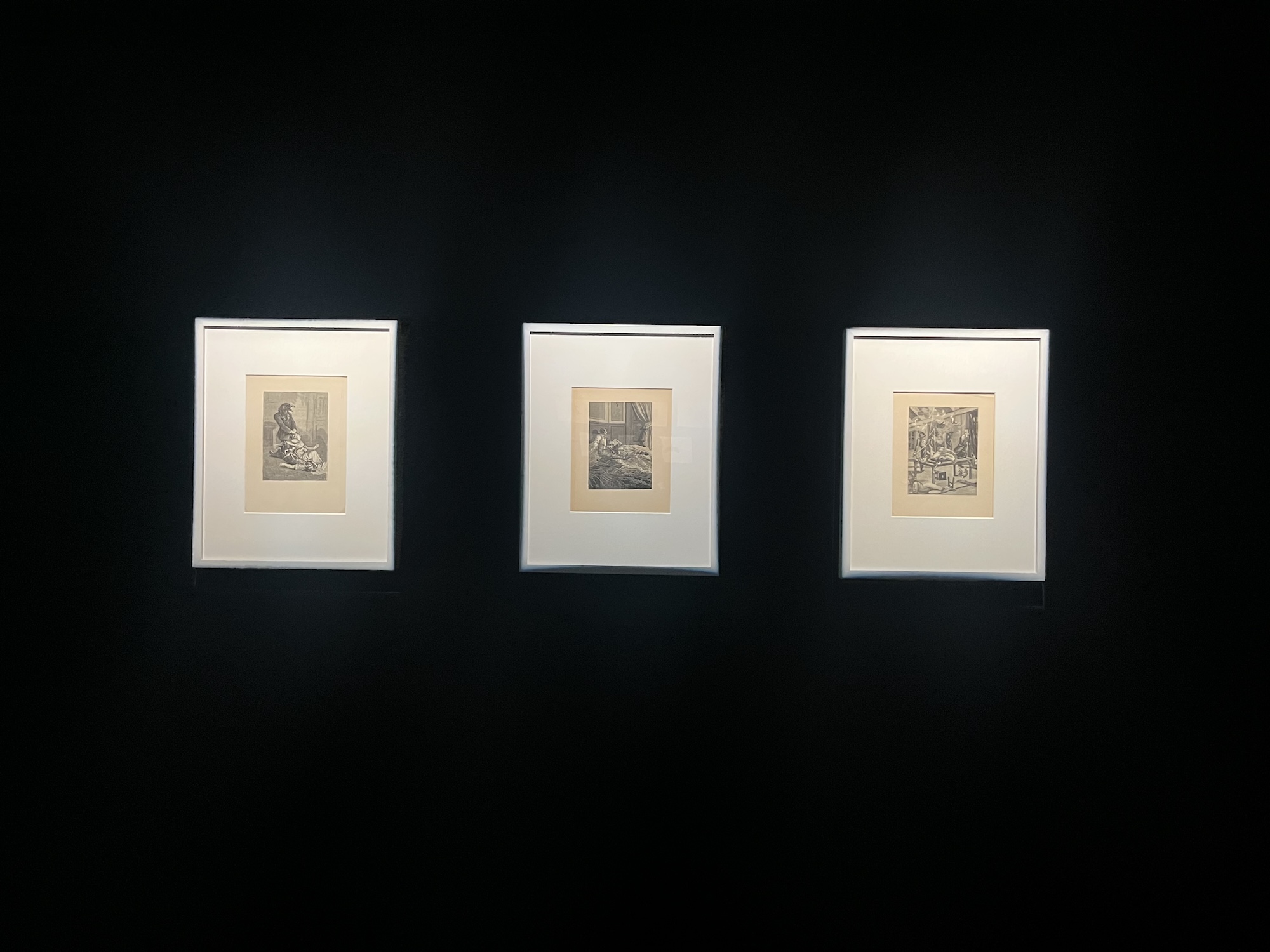 Installation view of three white frames hanging on a black wall, each containing a greyscale piece of art.