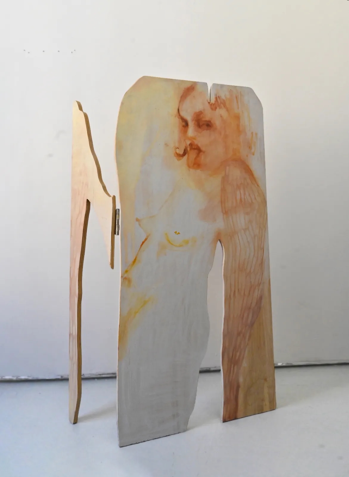 A three-dimensional sculptural work consisting of shaped plywood panels hinged together, painted with a loosely rendered figurative image of a nude figure in warm amber, rust, and white tones. The cutout silhouette allows the sculpture to stand freestanding.