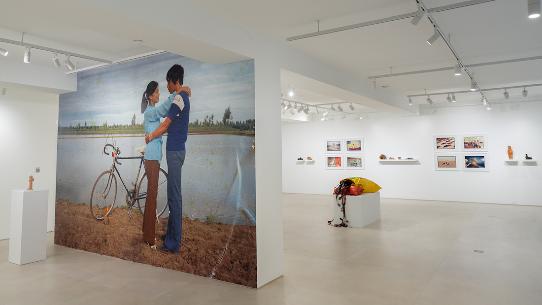 Gallery installation view featuring a large-scale photographic mural of an embracing couple beside a bicycle at a waterside, with the broader exhibition space visible beyond it, including framed photographs on the walls, small sculptures on shelves, and a deflated life raft on a white plinth.