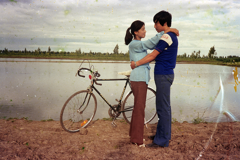 A young couple embraces beside a bicycle at the edge of a flooded field, overcast sky reflected in the water behind them. Vintage film photograph with visible grain and scratches.