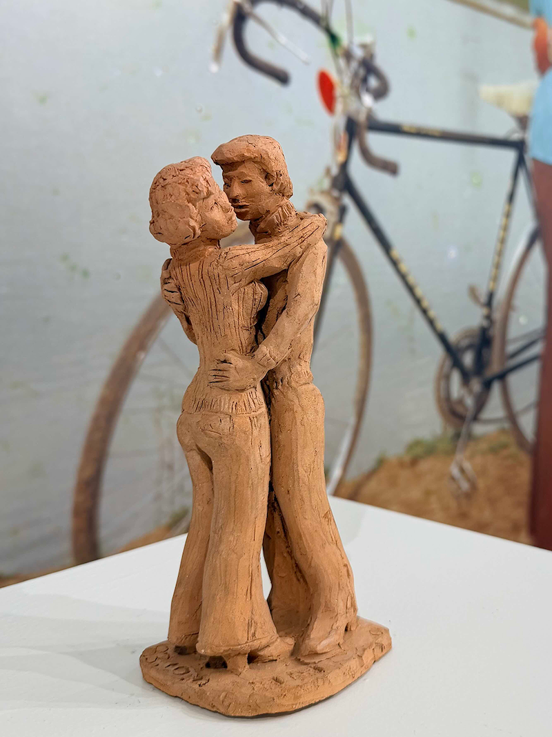 A terracotta clay sculpture of an embracing couple, positioned in front of a blurred photographic enlargement of the couple from Image 1 with a bicycle visible in the background.