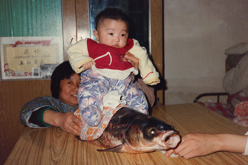 A grandmother holds a bundled infant upright on a table next to a large whole carp fish, smiling warmly. Vintage snapshot with a framed certificate visible on the wall behind them.