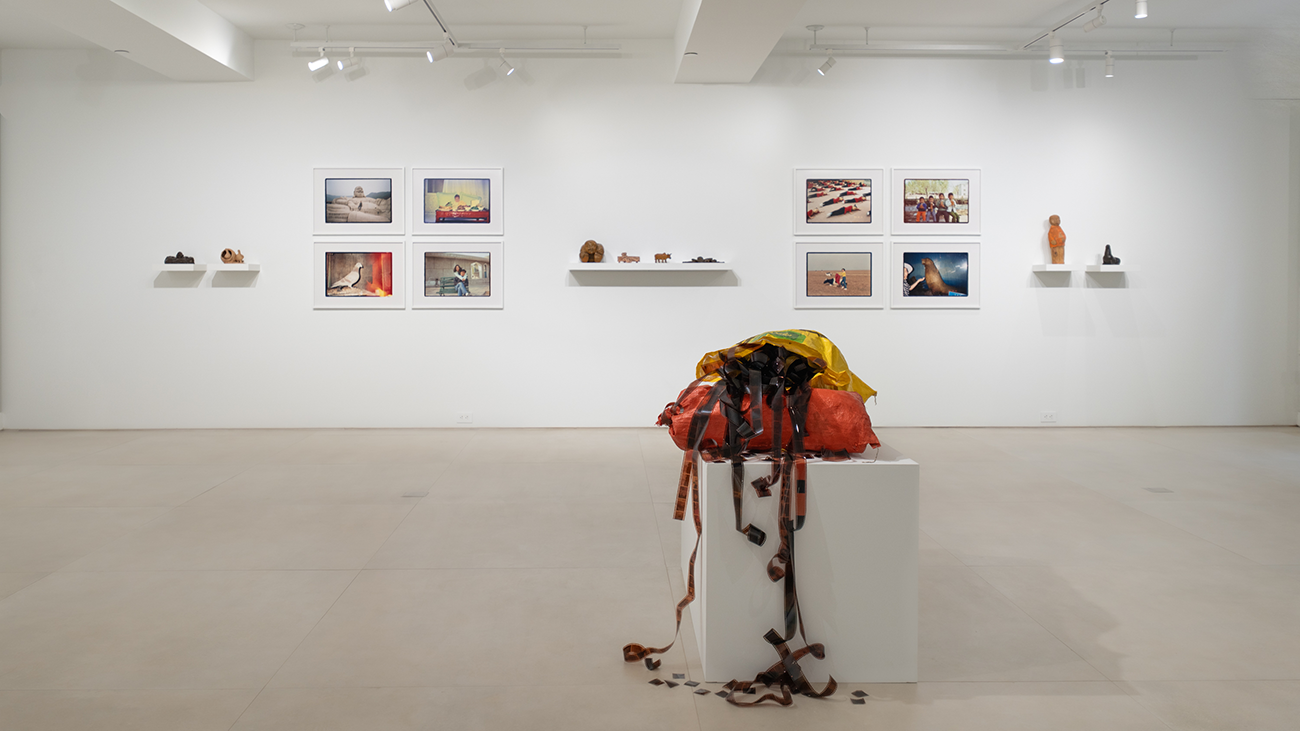 Gallery installation view with eight framed color photographs arranged in two groups on a white wall, small sculptural objects on shelves between them, and a deflated orange and yellow life raft displayed on a white plinth in the foreground.