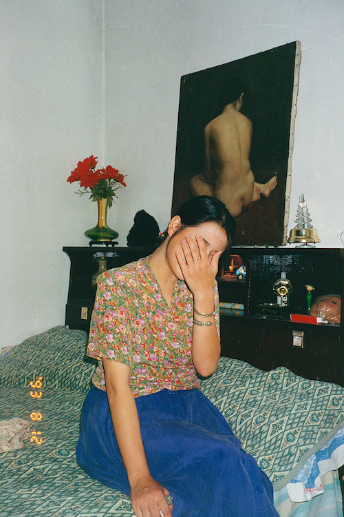 A person in a floral blouse and blue skirt sits on a bed with their face covered by one hand, a figurative painted canvas hanging on the wall behind them.