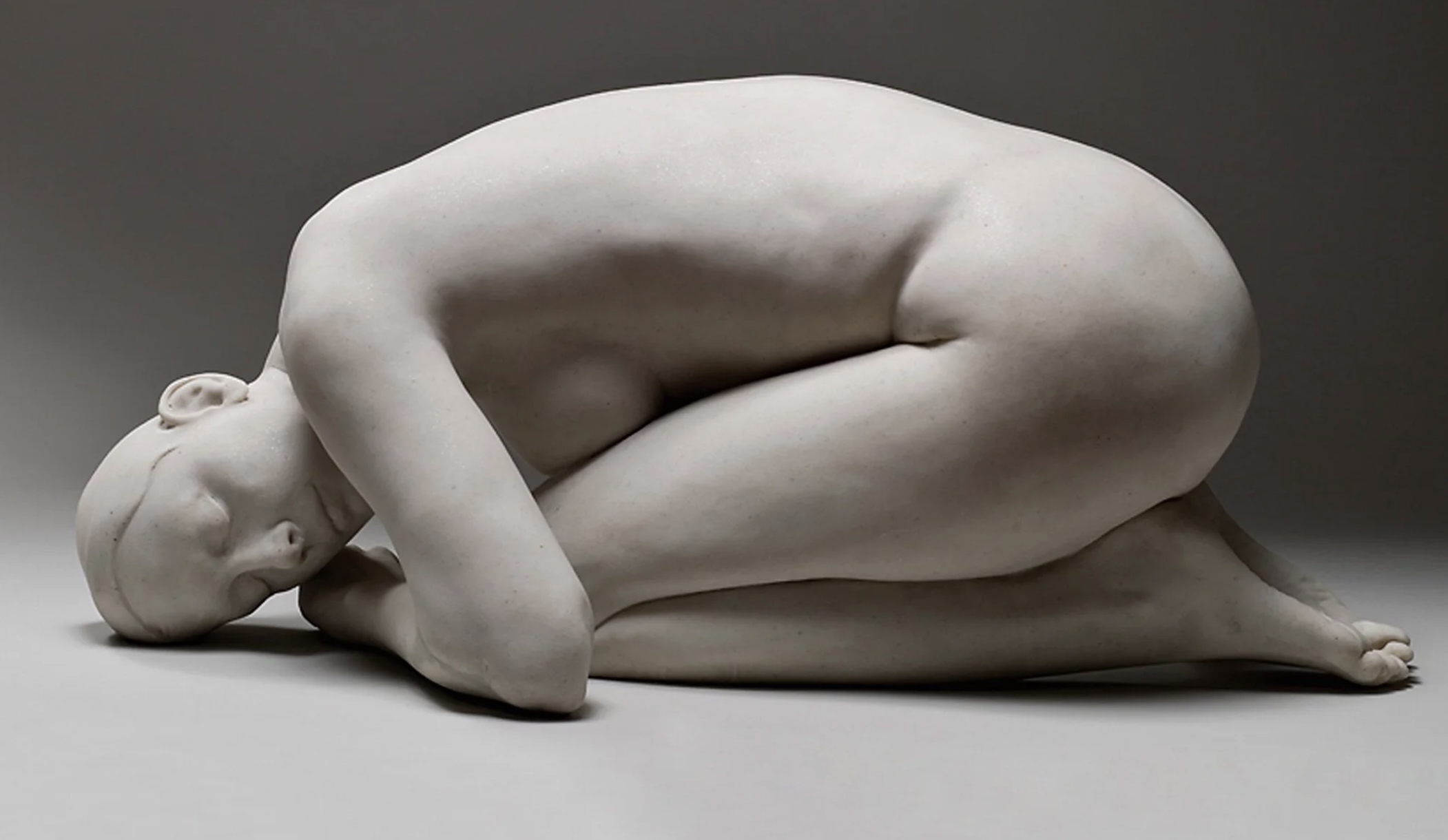 White marble sculpture of a human figure curled into a tight fetal position, lying on its side against a neutral gray background.