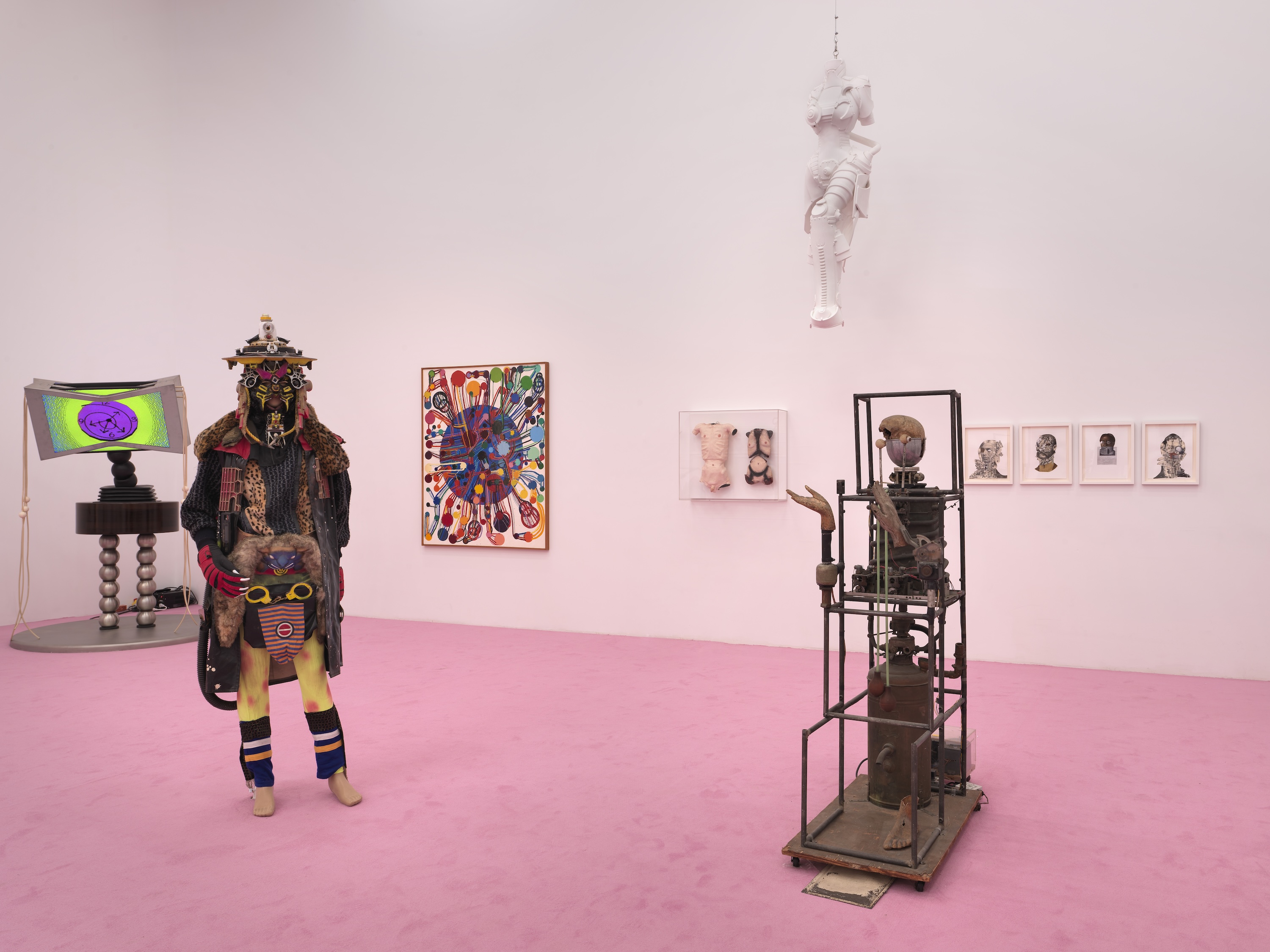Gallery room with a pink carpet featuring an elaborately costumed figure in layered animal pelts and an ornate headdress, alongside a mechanical tower sculpture and suspended white figure.
