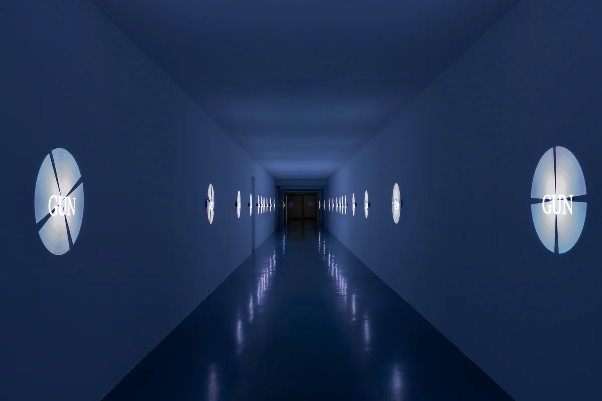 Installation view of a dark blue corridor with illuminated circular wall-mounted works displaying the word "GUN," receding into the distance.