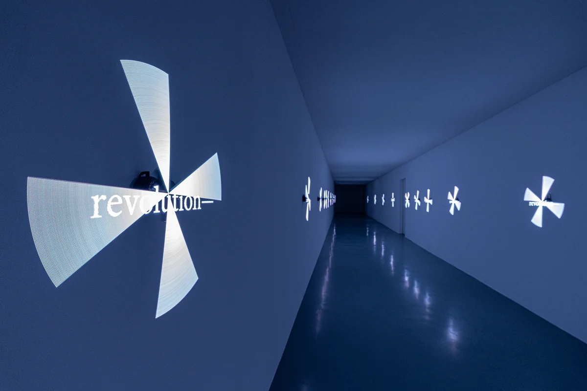 Installation view of a dark blue corridor with spinning pinwheel-like projections on the walls, the nearest reading "revolution–."