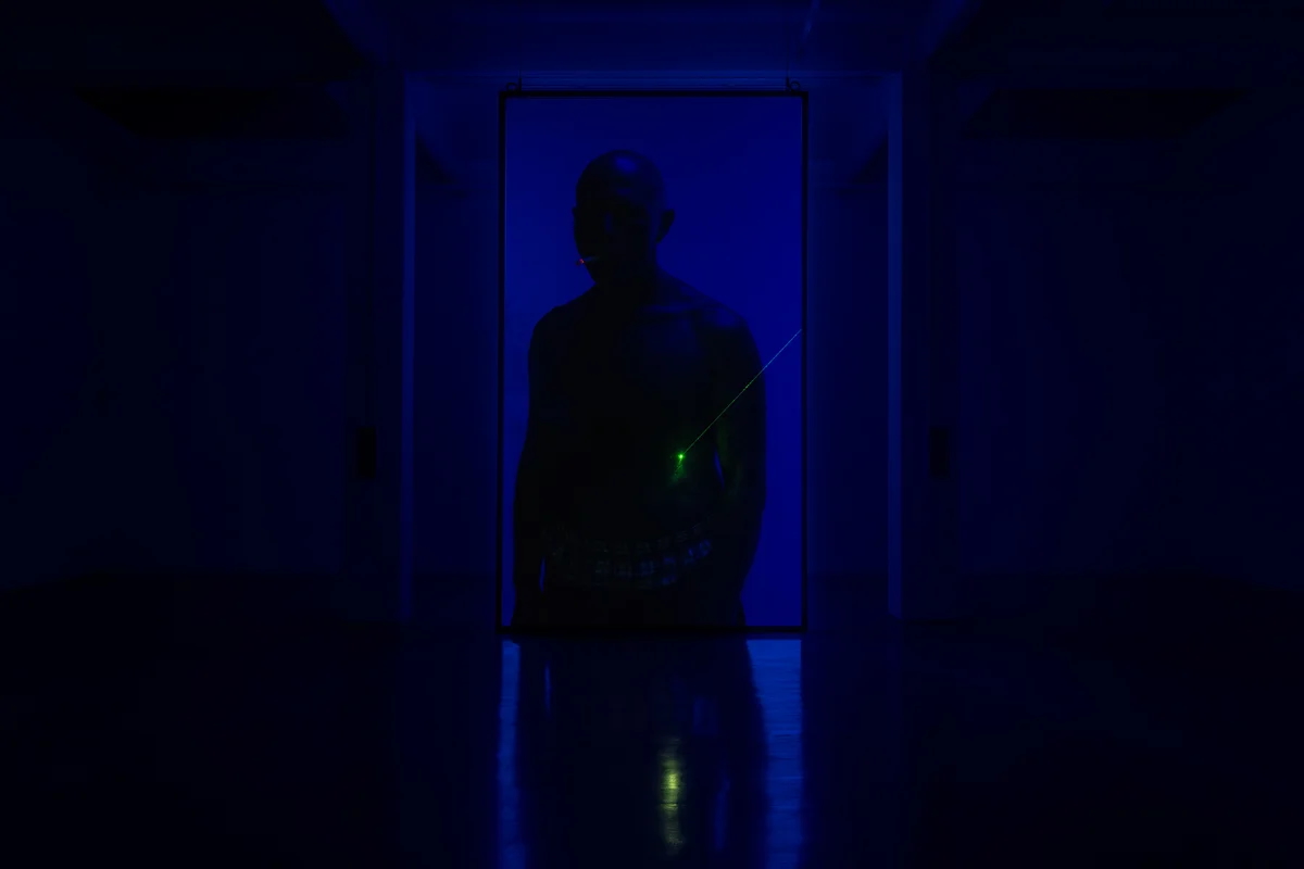 A silhouetted figure stands backlit in blue light at the end of a dark corridor, a green laser line crossing their torso.