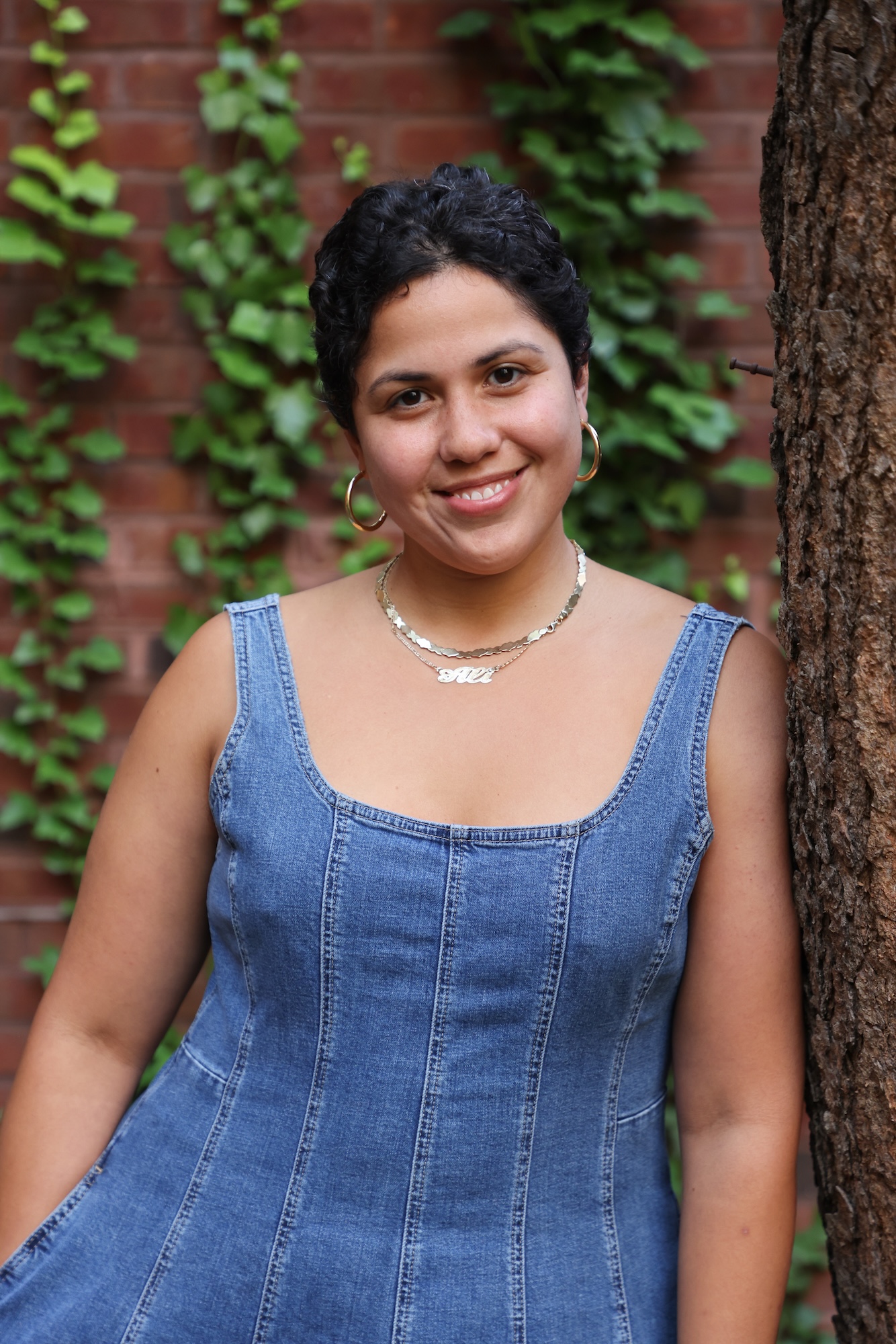 Portrait of a smiling person in a denim dress standing beside a tree.