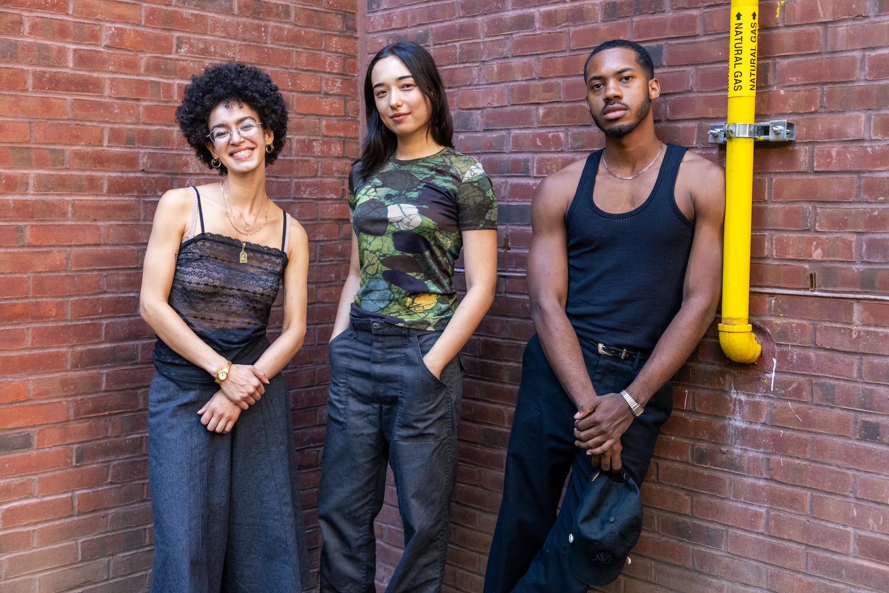 Three young adults pose against a brick wall in an outdoor courtyard.