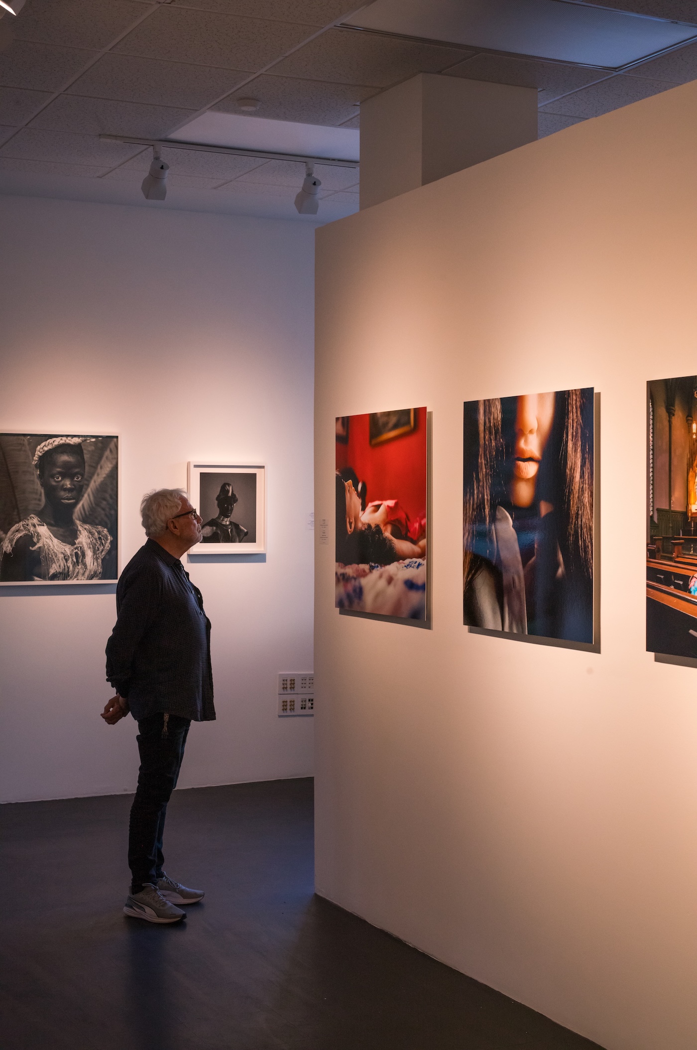 A gallery visitor looks up at photographs mounted on a white wall in a dimly lit exhibition space.
