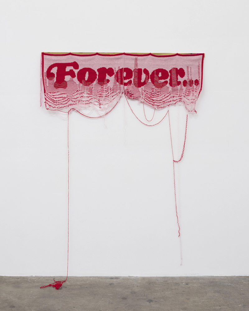 Knitted textile wall piece spelling "Forever…" in red letters on a lighter red ground, with loose unraveling threads hanging down to the floor.