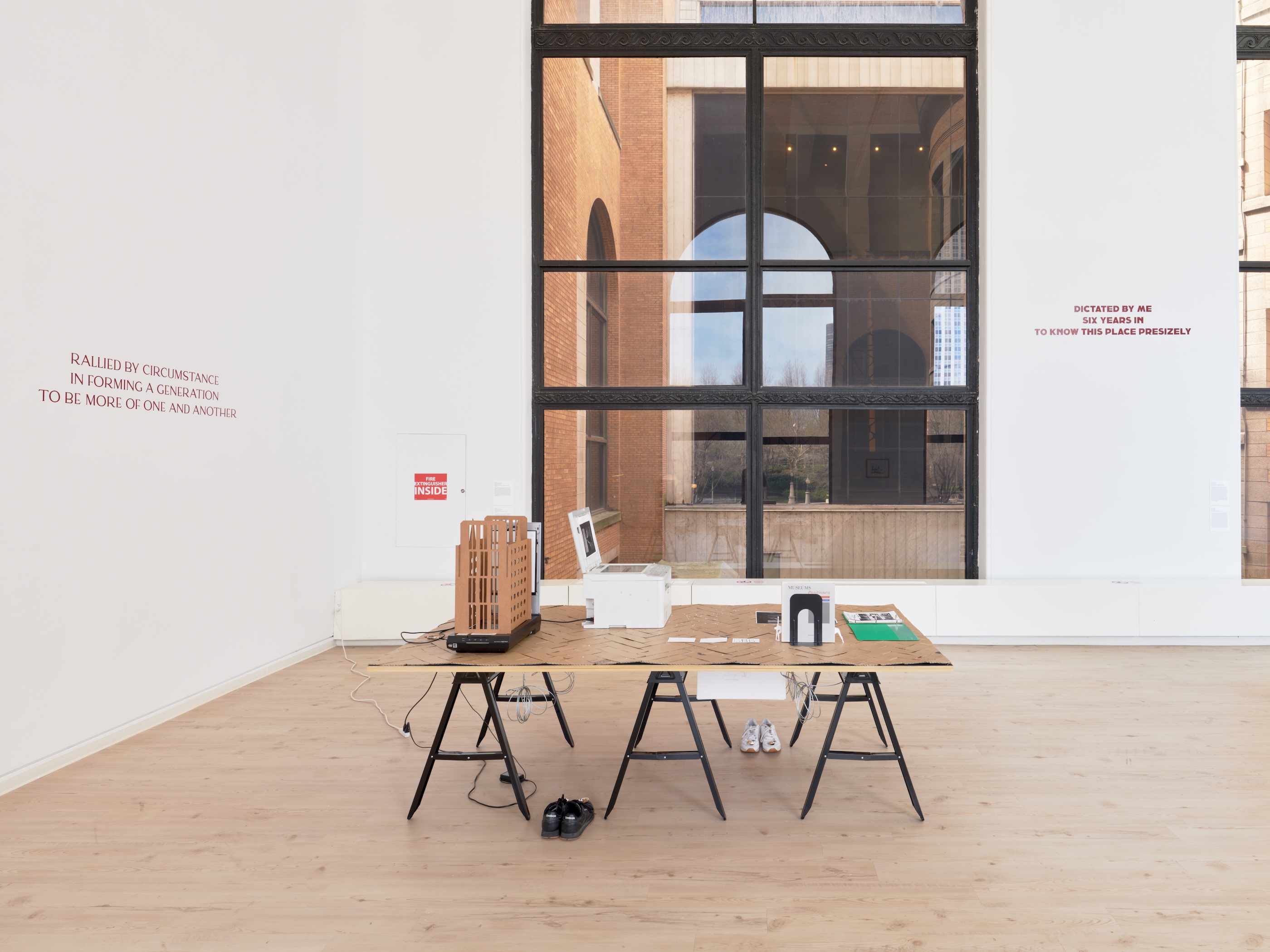 Gallery installation view showing a large trestle table covered in cardboard, holding a cardboard architectural model, a printer, and various objects, with sneakers placed underneath. Two walls bear maroon text in a serif font, with floor-to-ceiling windows revealing an adjacent brick building.