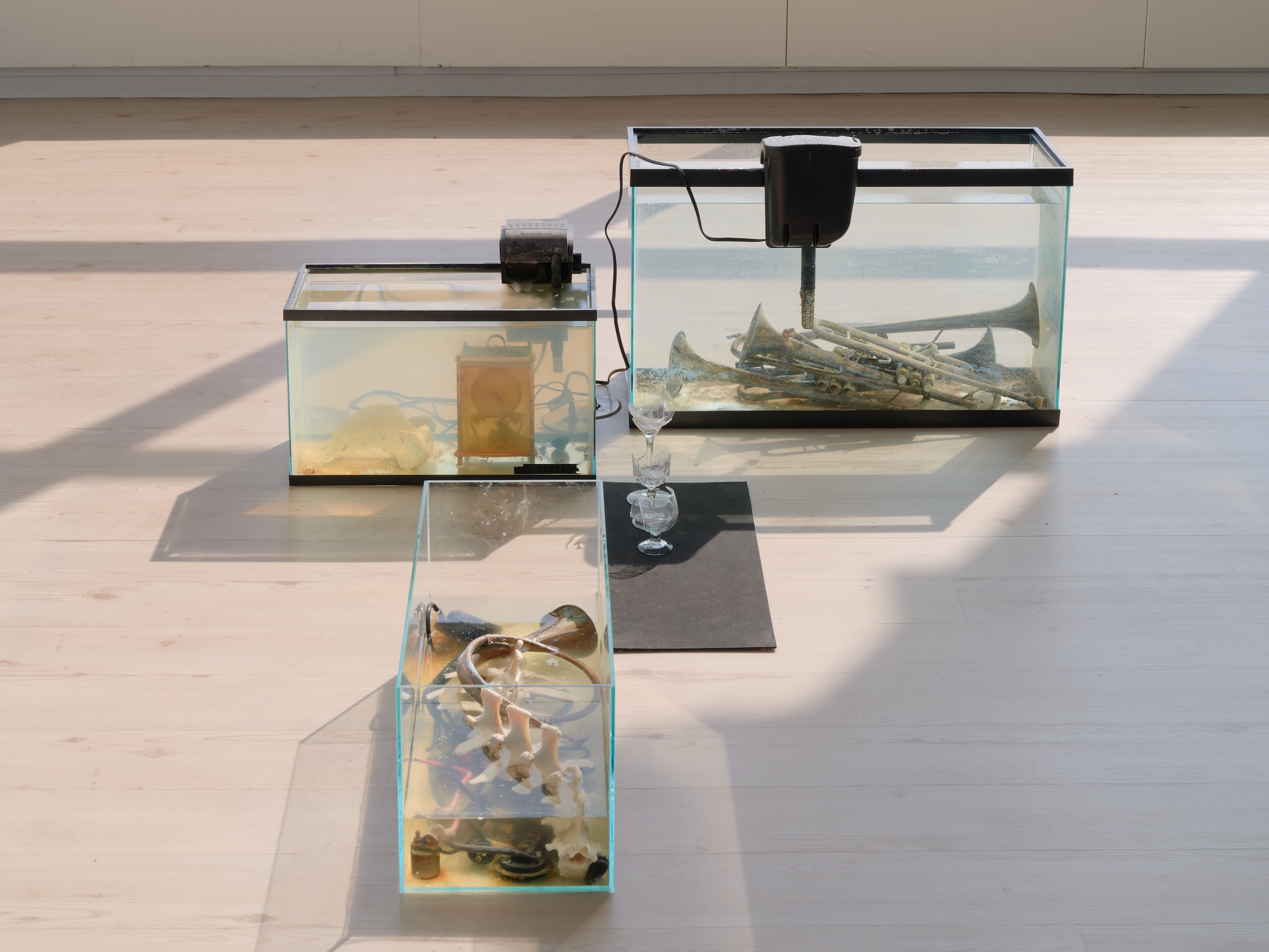 Three glass tanks filled with liquid arranged on a wood floor, containing submerged objects including tarnished brass trumpets, bones, electronic components, and a glowing orange device. A pair of stacked crystal wine glasses sits on a dark mat nearby.