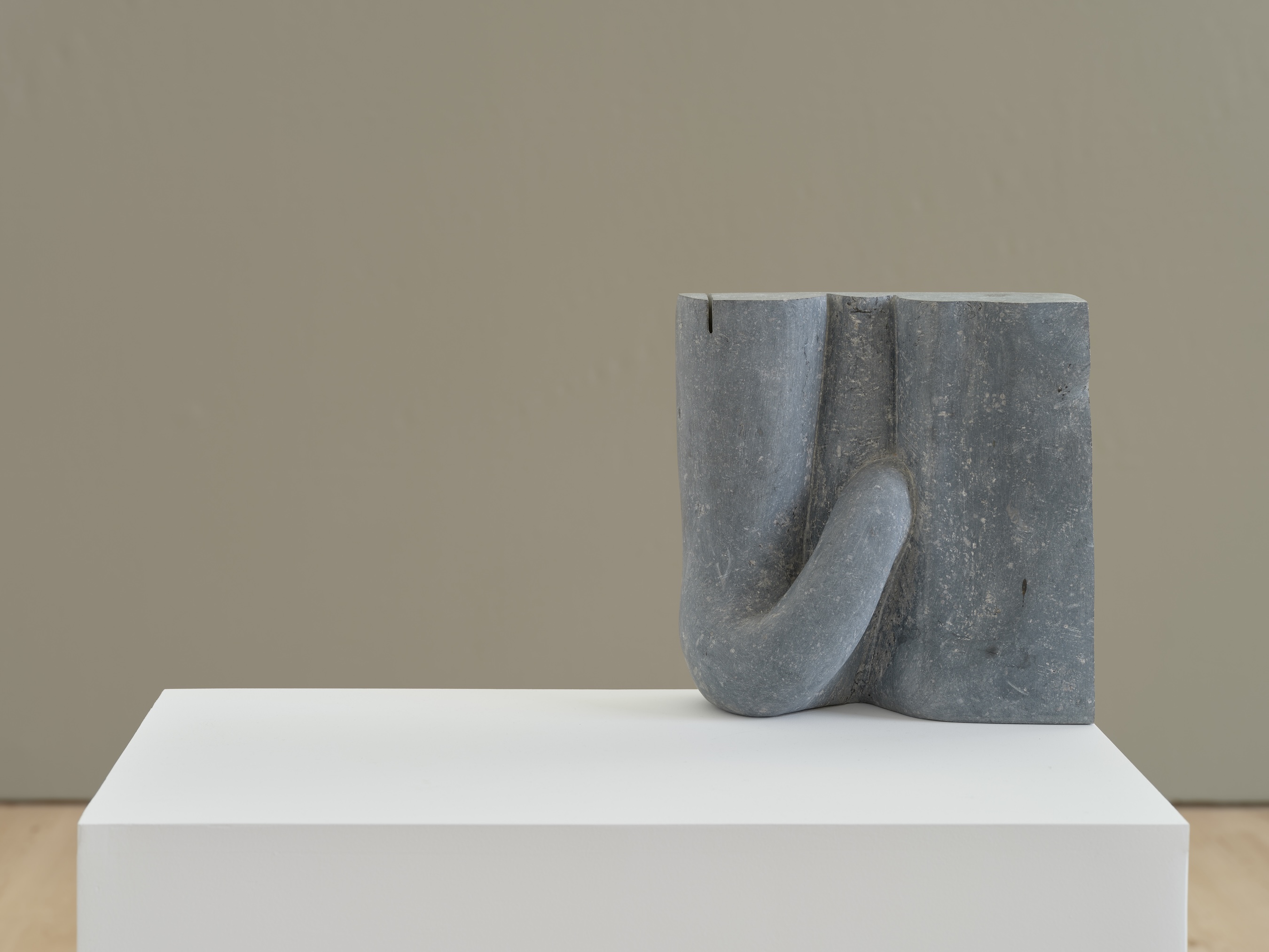 A smooth gray stone sculpture with an organic, looping form resembling a folded or knotted shape, displayed on a white pedestal against a tan wall.