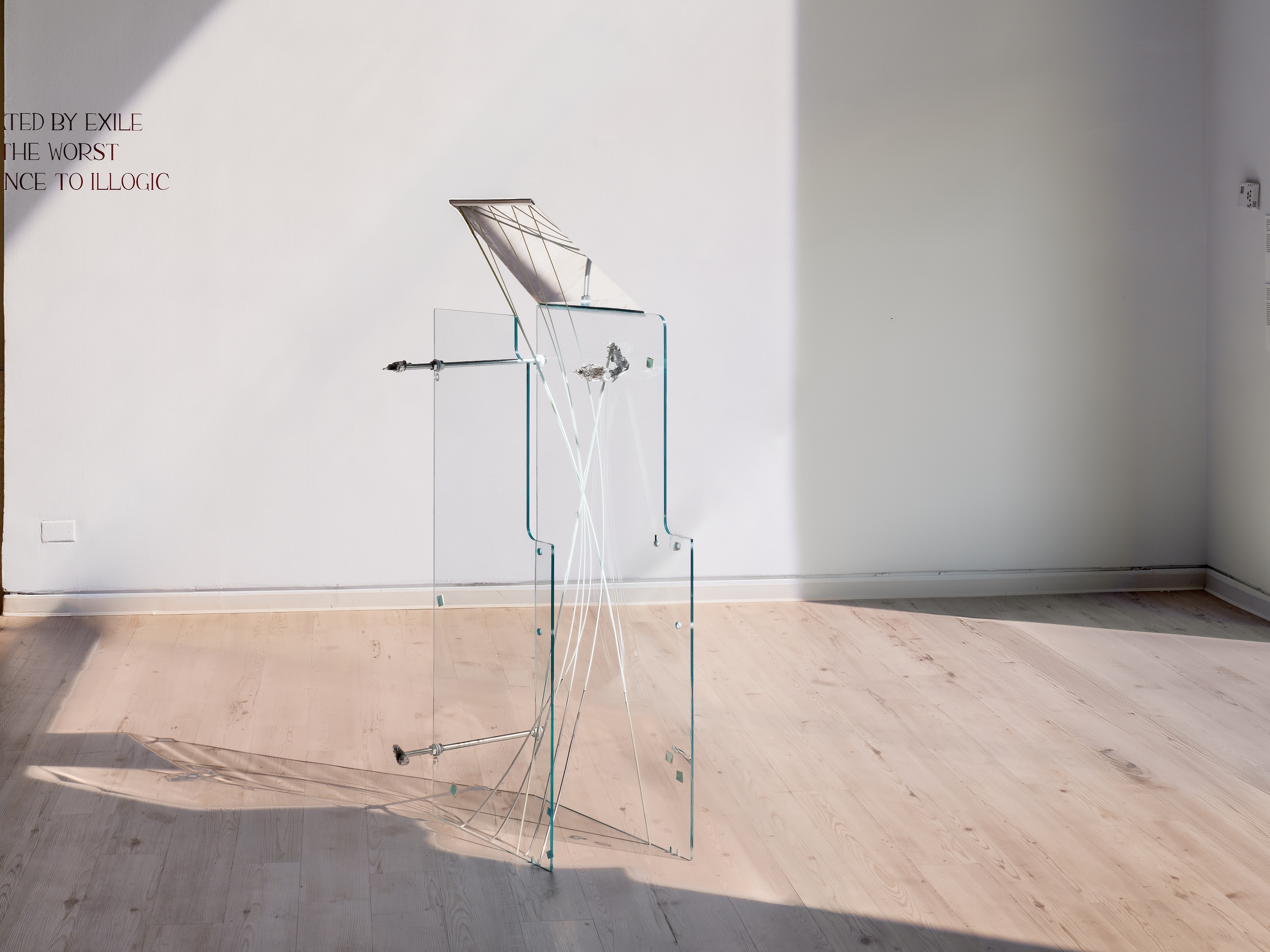A sculptural installation of sheet glass panels strung with taut white cords, topped by an angled mirrored or metallic panel, standing on a wood floor.