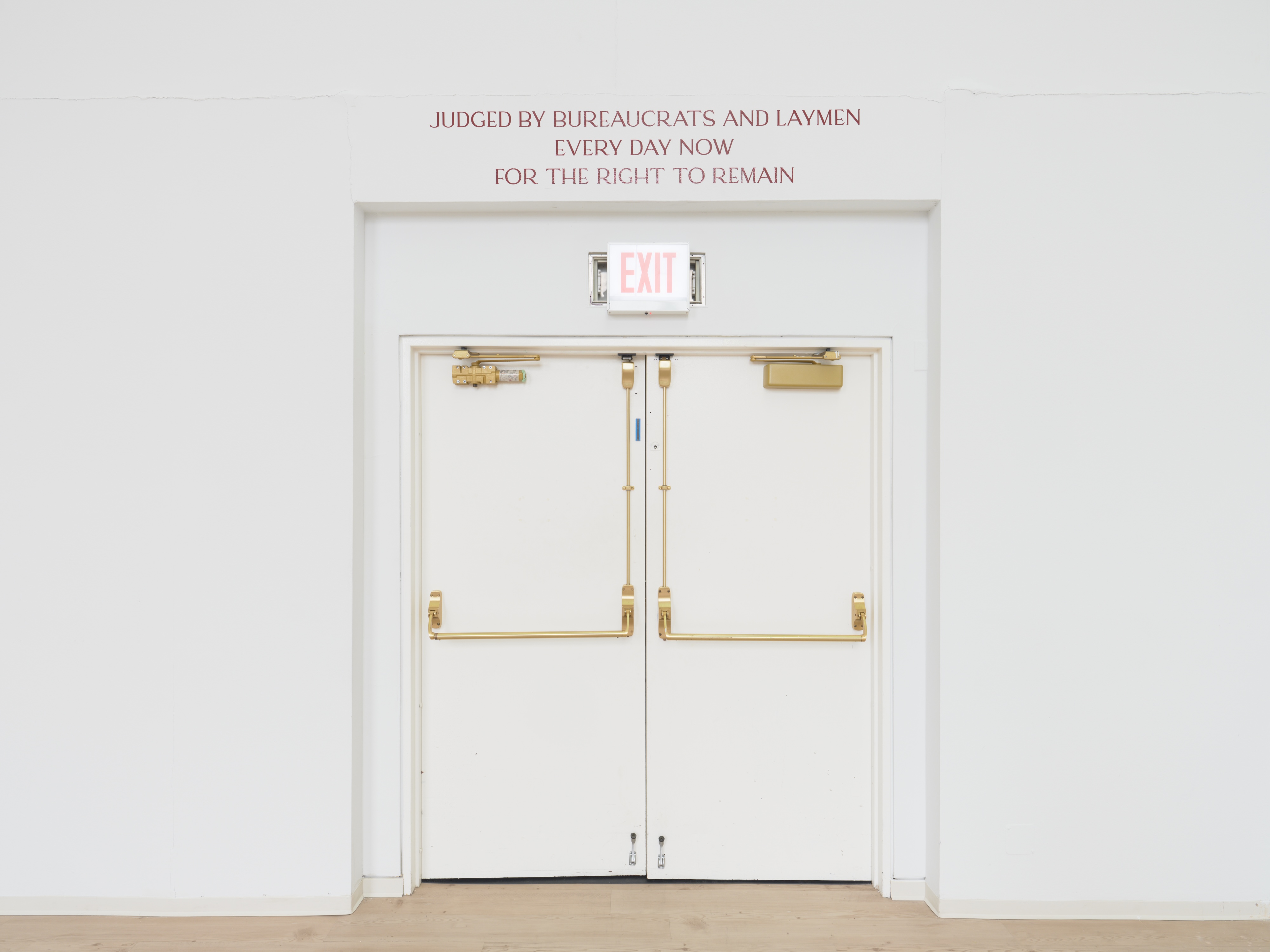 A white double exit door with brass push bars and an illuminated "EXIT" sign above, set into a white wall. Maroon serif text above the doorframe reads "JUDGED BY BUREAUCRATS AND LAYMEN EVERY DAY NOW FOR THE RIGHT TO REMAIN."