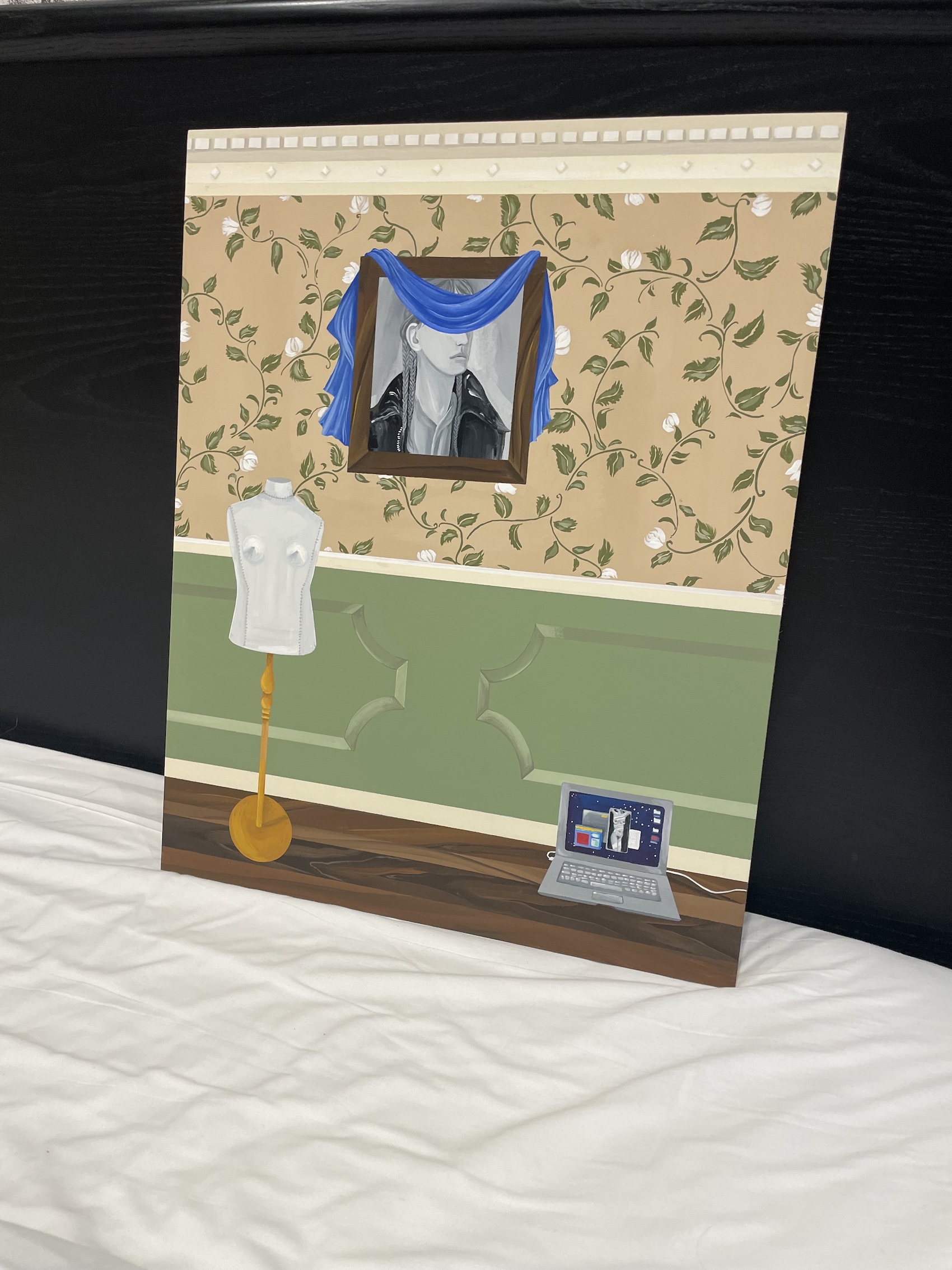 A painting propped against a dark wall, showing an interior scene with floral wallpaper above a green wainscoted wall. A framed black-and-white portrait of a person in a leather jacket with braided hair hangs draped with a blue scarf, beside a white dress form on a stand and an open laptop on the floor displaying a desktop with icons.