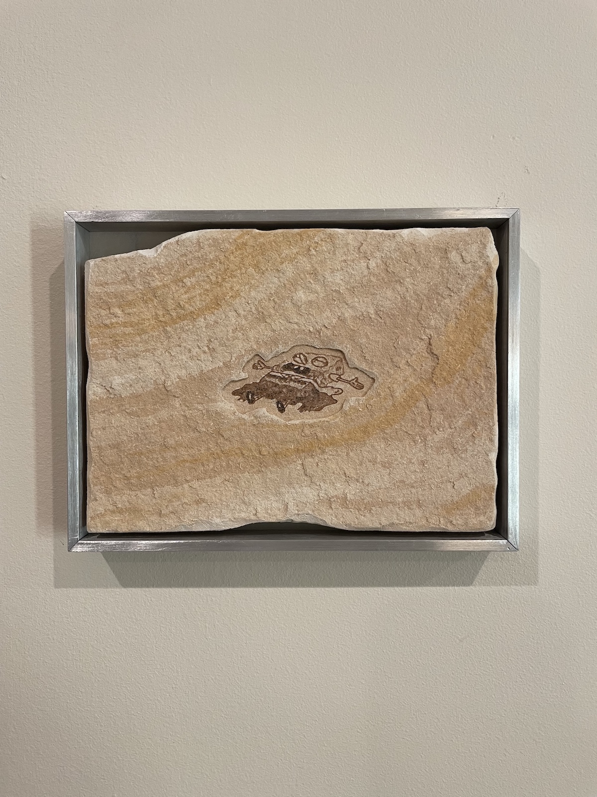 A rectangular slab of beige sandstone with natural striated patterns, mounted in a metal frame, featuring a dark fossil impression at its center resembling small interlocking shapes or figures in the form of the character SpongeBob.