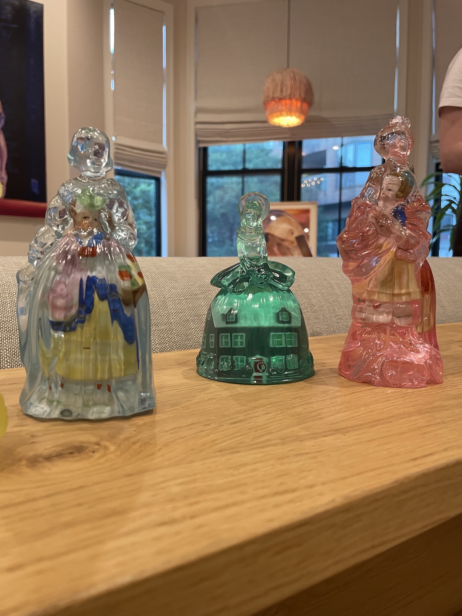 Three antique pressed-glass figural cologne or candy containers in clear, green, and pink glass, shaped as Victorian-dressed women and a house-shaped bell, displayed on a wooden table in front of a window.