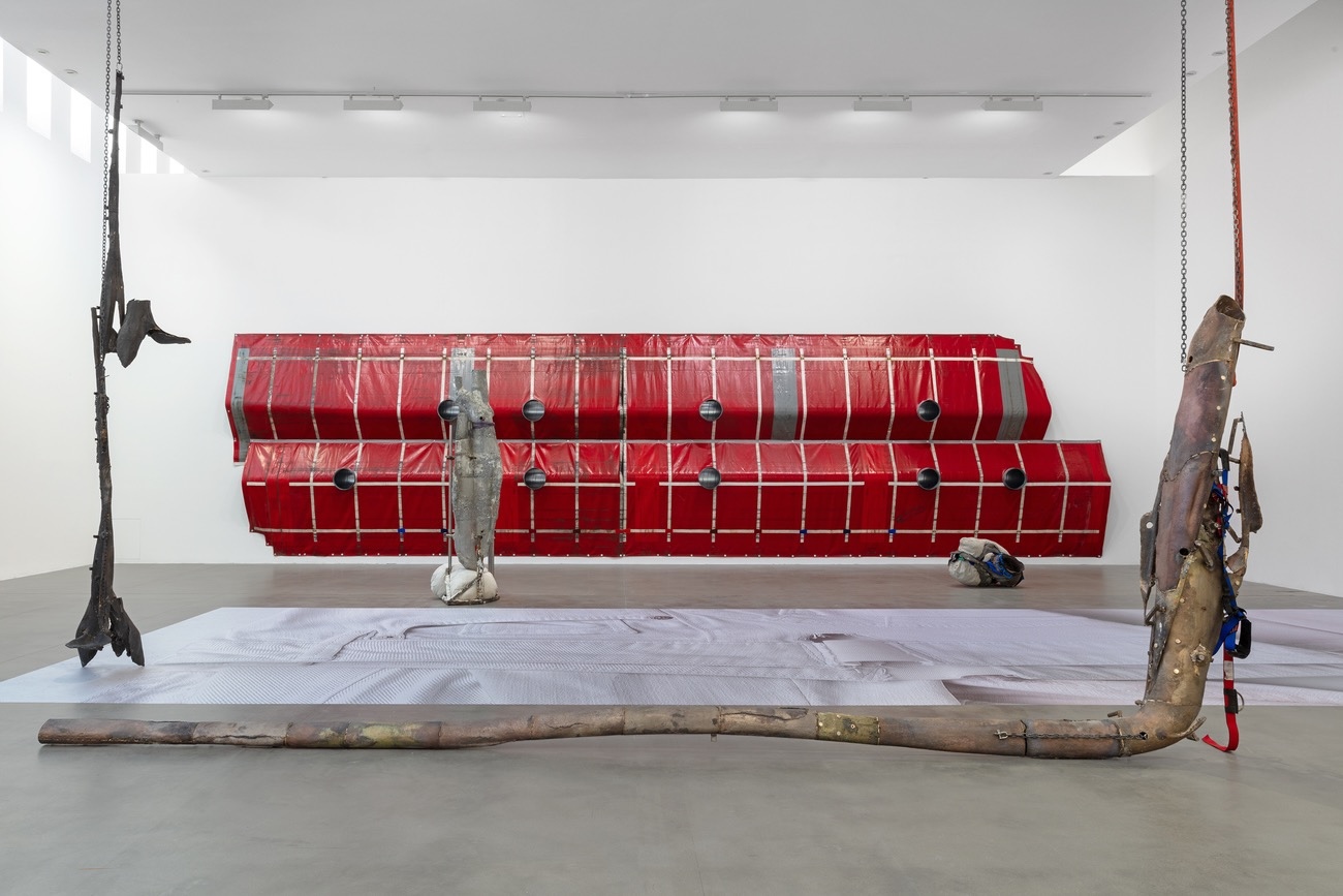 Gallery installation featuring a large red modular wall-mounted structure with circular openings, flanked by two hanging metal sculptural forms and a curved wooden branch on the floor.