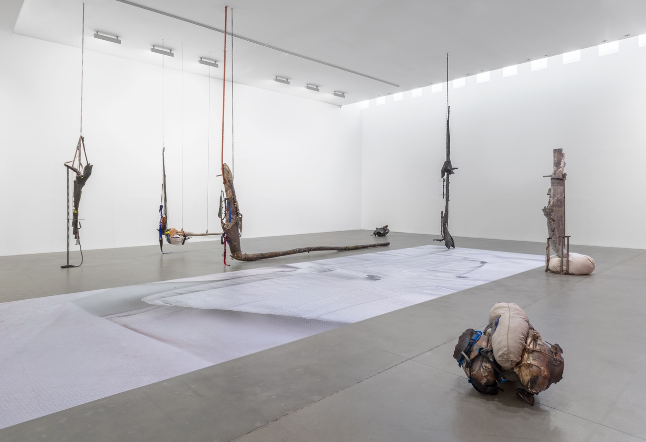 Wide-angle installation view showing multiple hanging sculptural assemblages made from found materials — branches, metal, and fabric — suspended above a large white textile on the gallery floor, with a bundled organic form in the foreground.