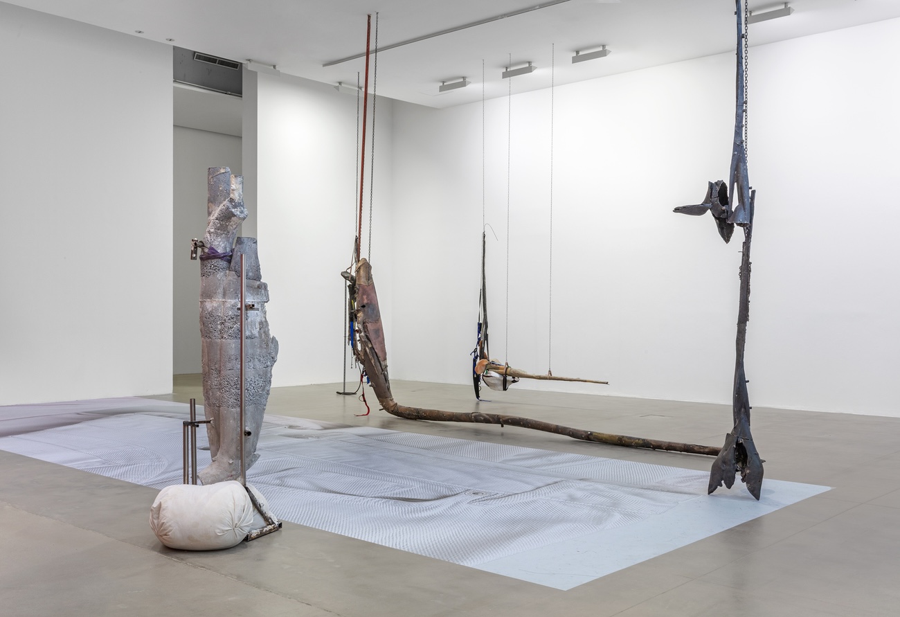 Installation view of sculptural works in a white gallery space, including a weathered column-like form, suspended metal and wood assemblages hanging from wires and chains, and a pale textile laid flat on the floor.