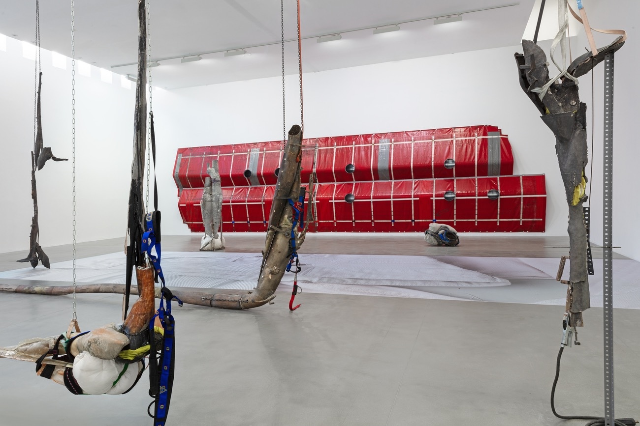 Installation view with several suspended assemblages of metal, wood, and strapping in the foreground, a large red paneled wall piece in the background, and a bare tree trunk mounted vertically on the right.