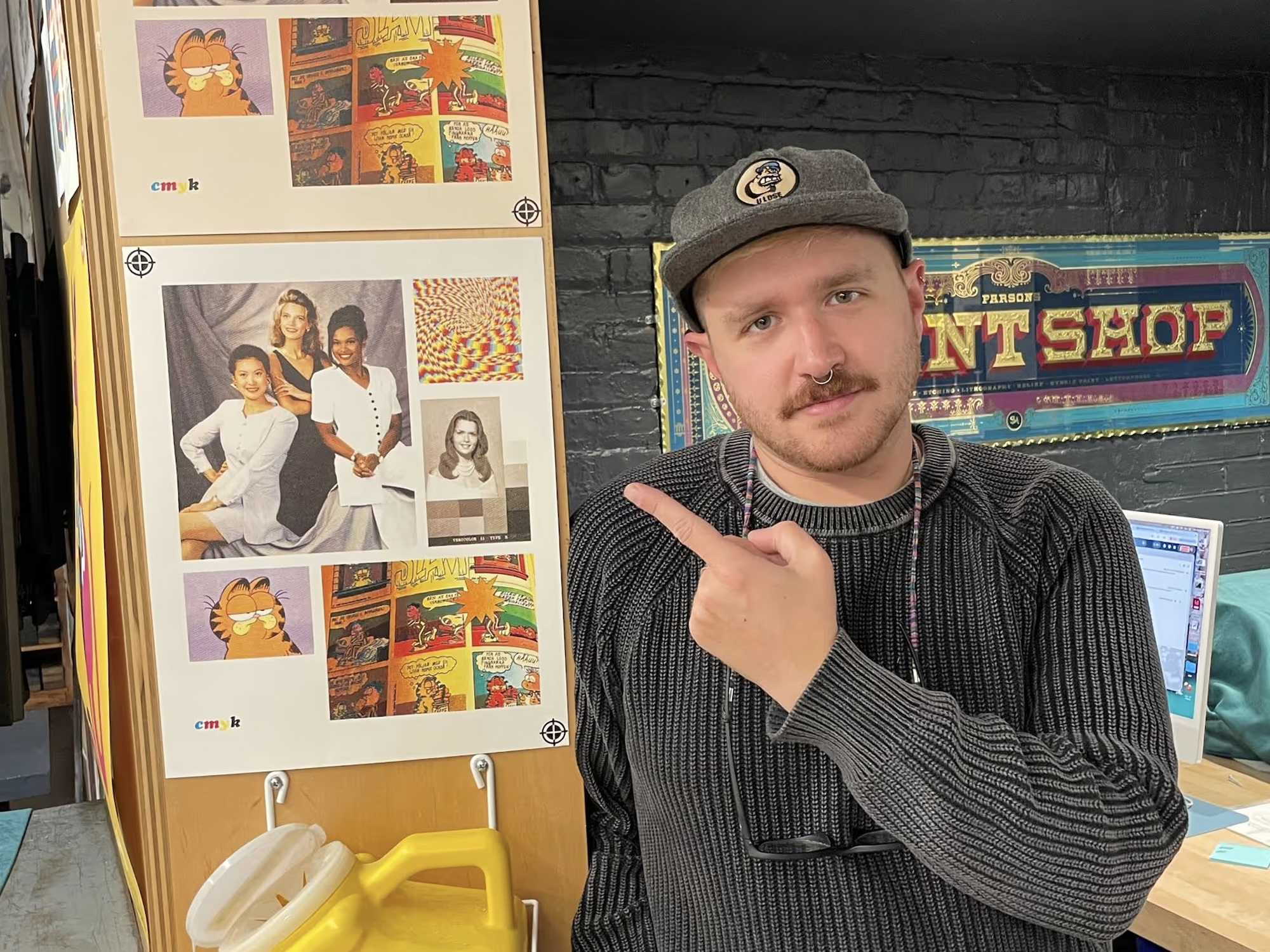 A man in a gray cap and knit sweater points at a CMYK test print board beside him, standing in front of a decorative 'Print Shop' sign.
