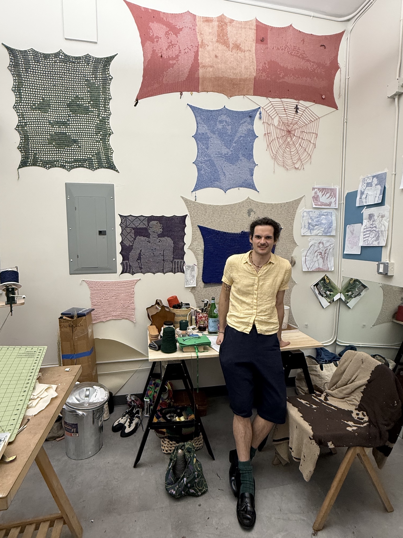 A smiling person in a yellow shirt leans against a worktable in an art studio, surrounded by large colorful crocheted textile works pinned to the walls.