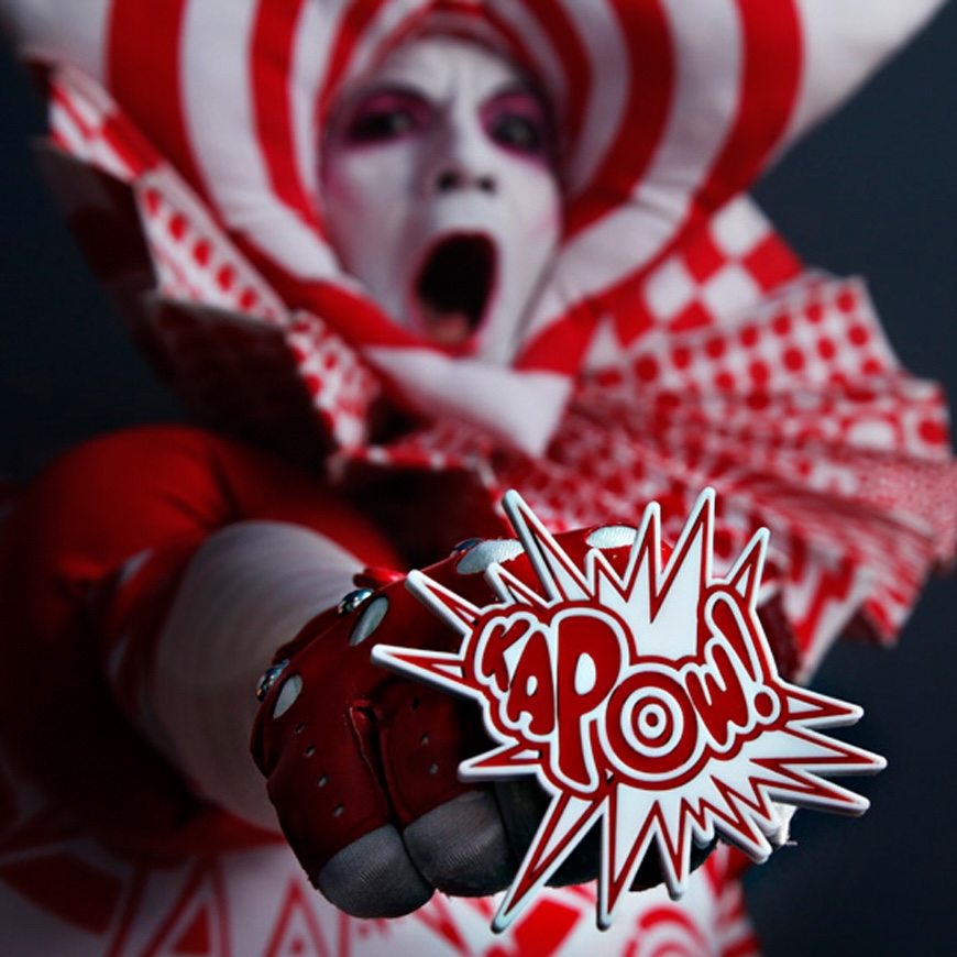 A performer in red-and-white jester costume and white face paint thrusts a fist toward the camera holding a comic-book-style 'KAPOW!' sign.