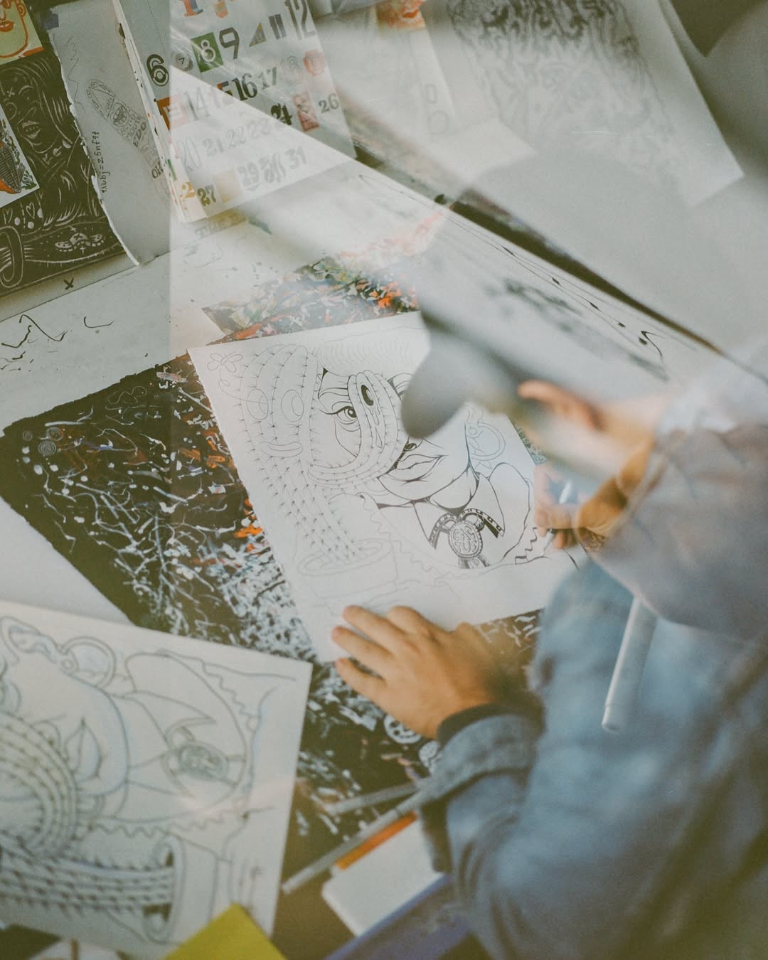 n artist's hands sorting through illustrated drawings and prints spread across a worktable, shot from above with a dreamy double-exposure effect.