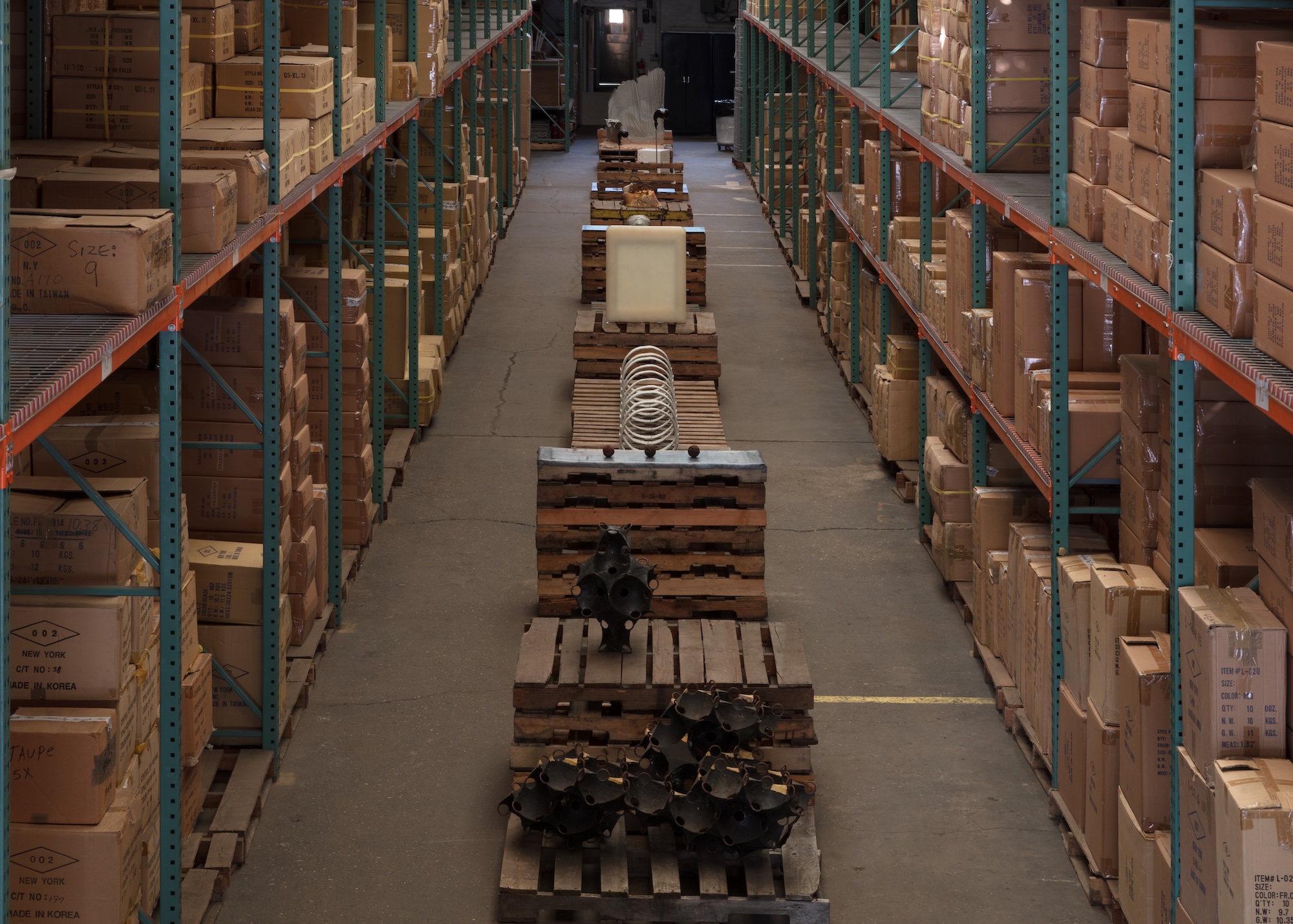 A warehouse aisle flanked by towering shelves of cardboard boxes, with a series of sculptural objects displayed on stacked wooden pallets receding into the distance.