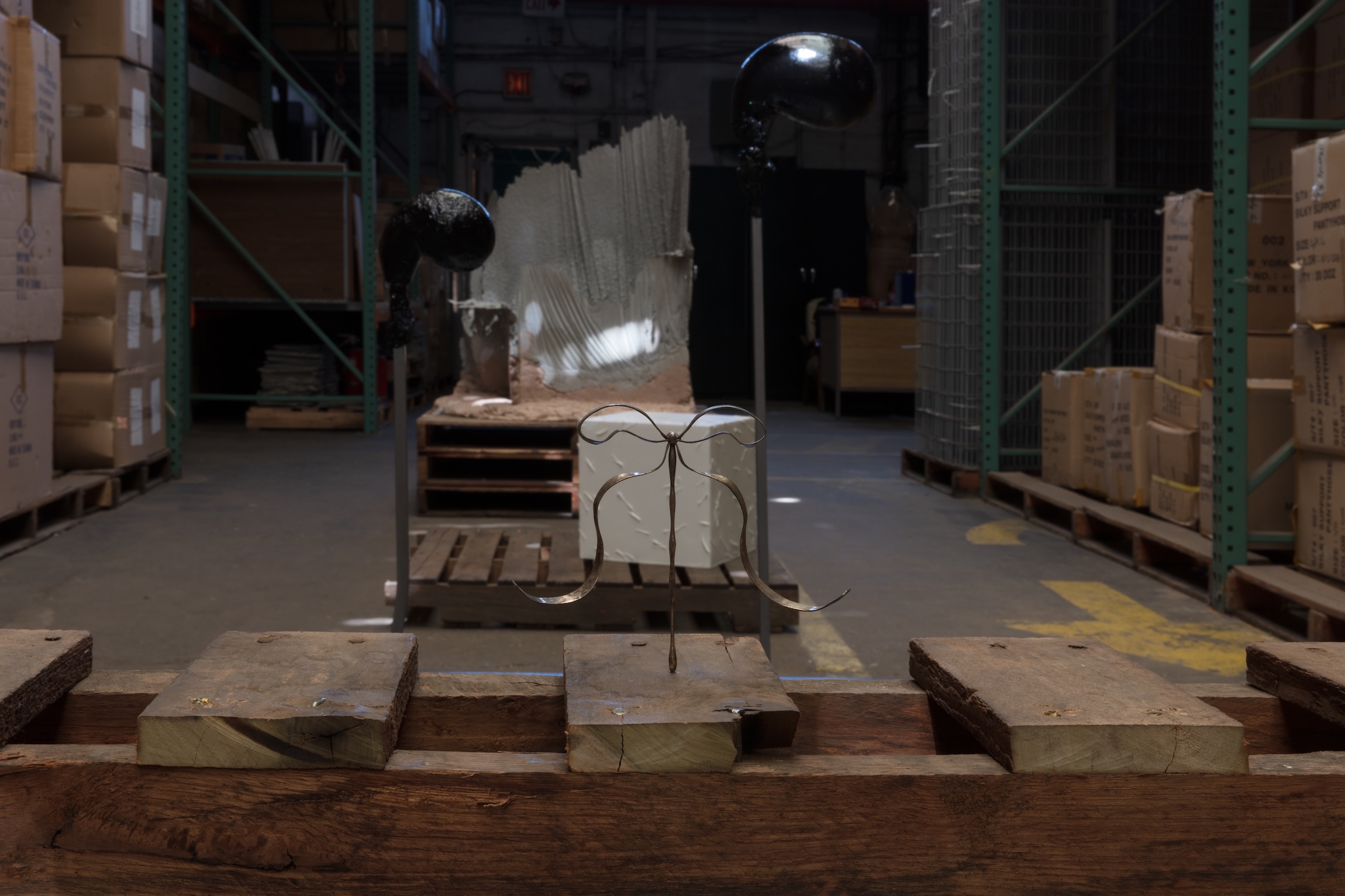 A delicate metal wire sculpture with curved legs sits on a pallet in a dimly lit warehouse aisle, with two black spheres on poles and a large white textured form visible in the background.