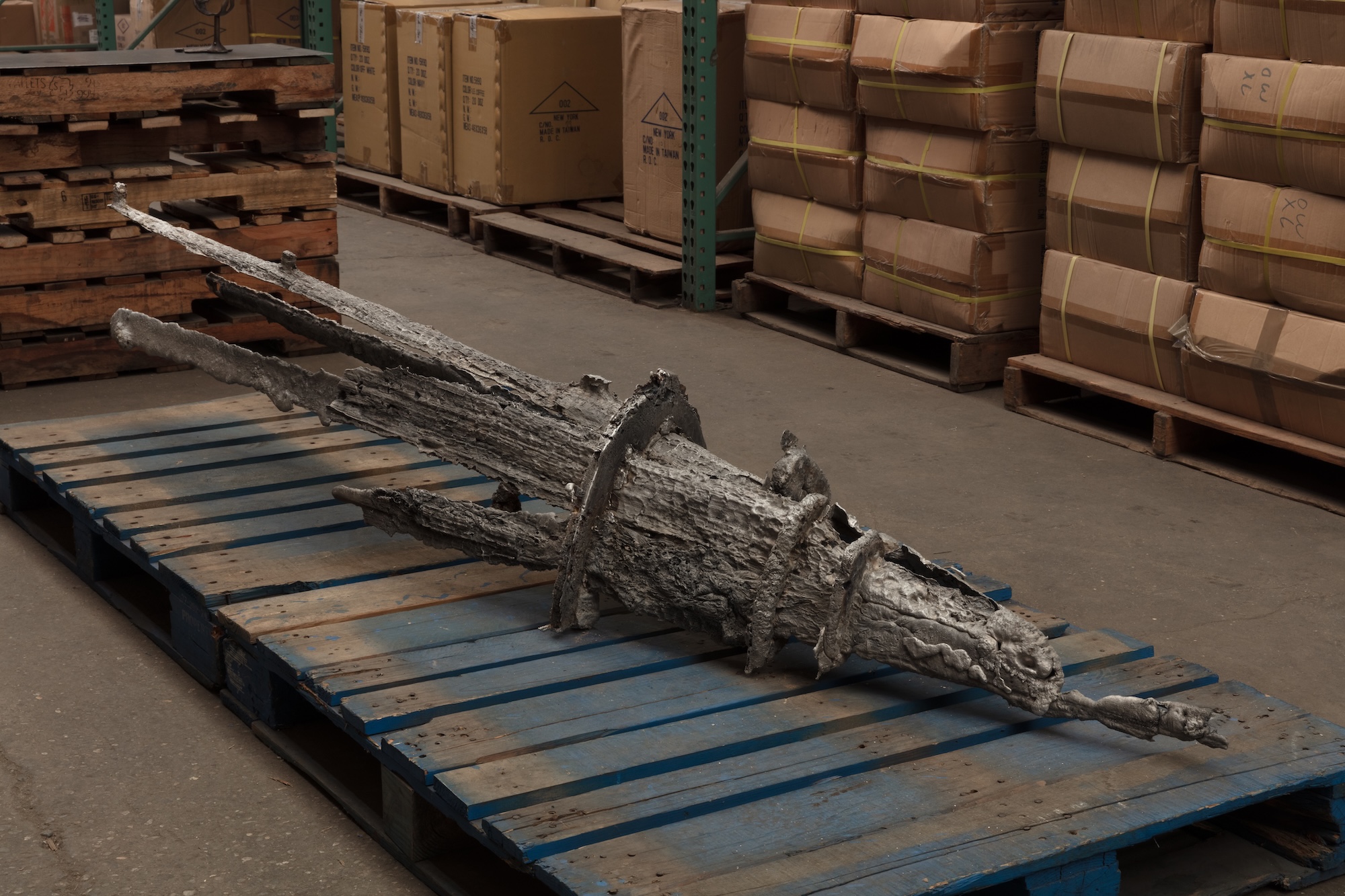 A large rough-textured cast metal sculpture resembling a weathered log or branch rests horizontally on a blue wooden pallet in a warehouse.