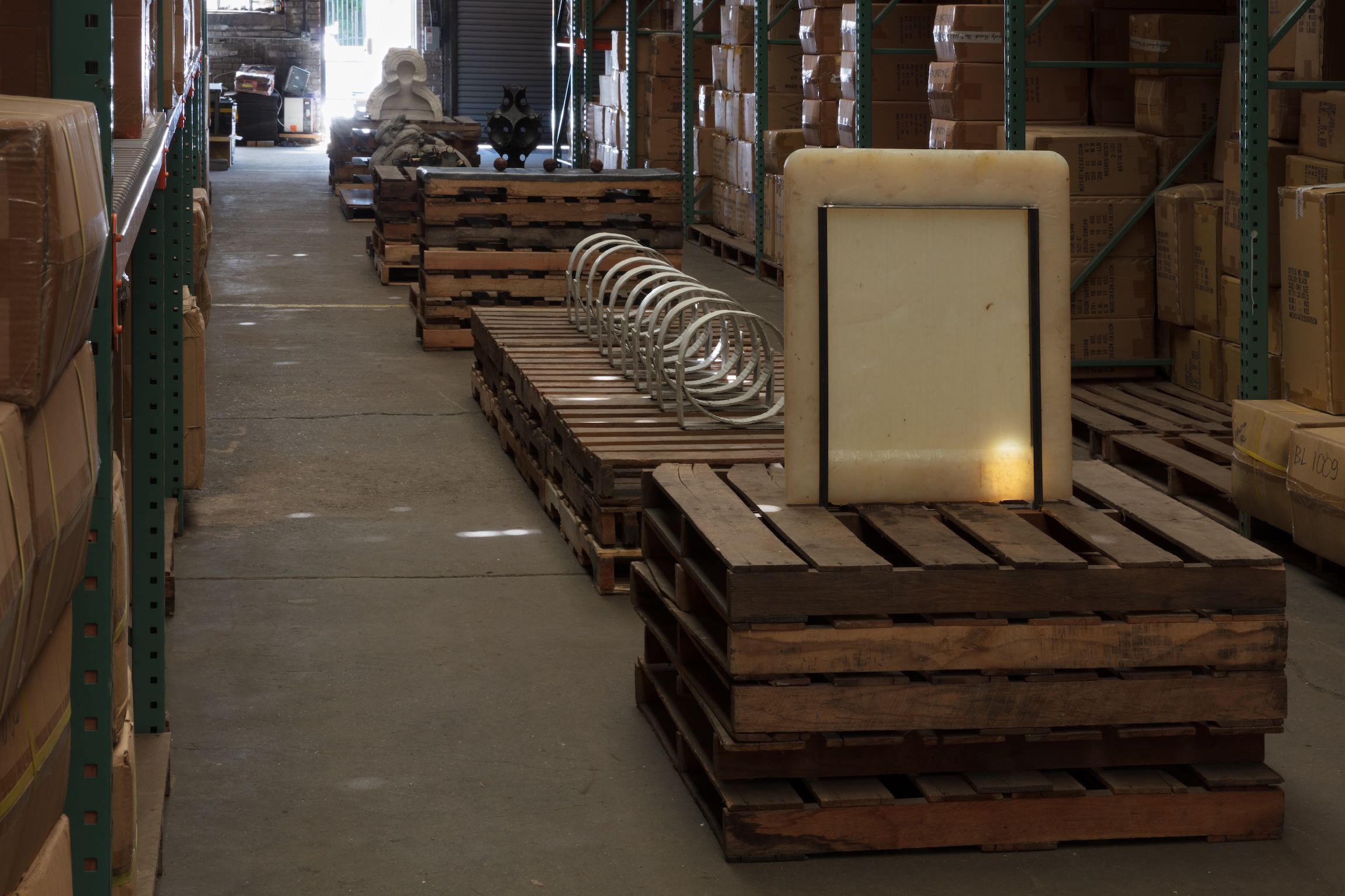 Close view of a warehouse aisle showing a glowing translucent white panel and a coiled metal spring displayed on stacked wooden pallets, with cardboard boxes on shelving behind.