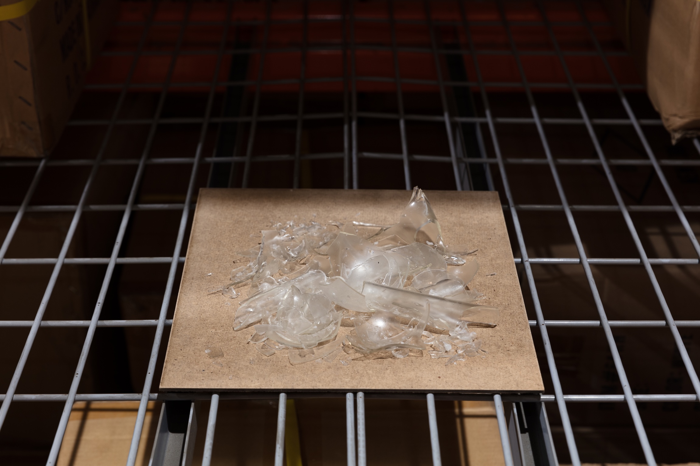 A pile of shattered clear glass objects sits on a square board atop a wire storage shelf, viewed from above.