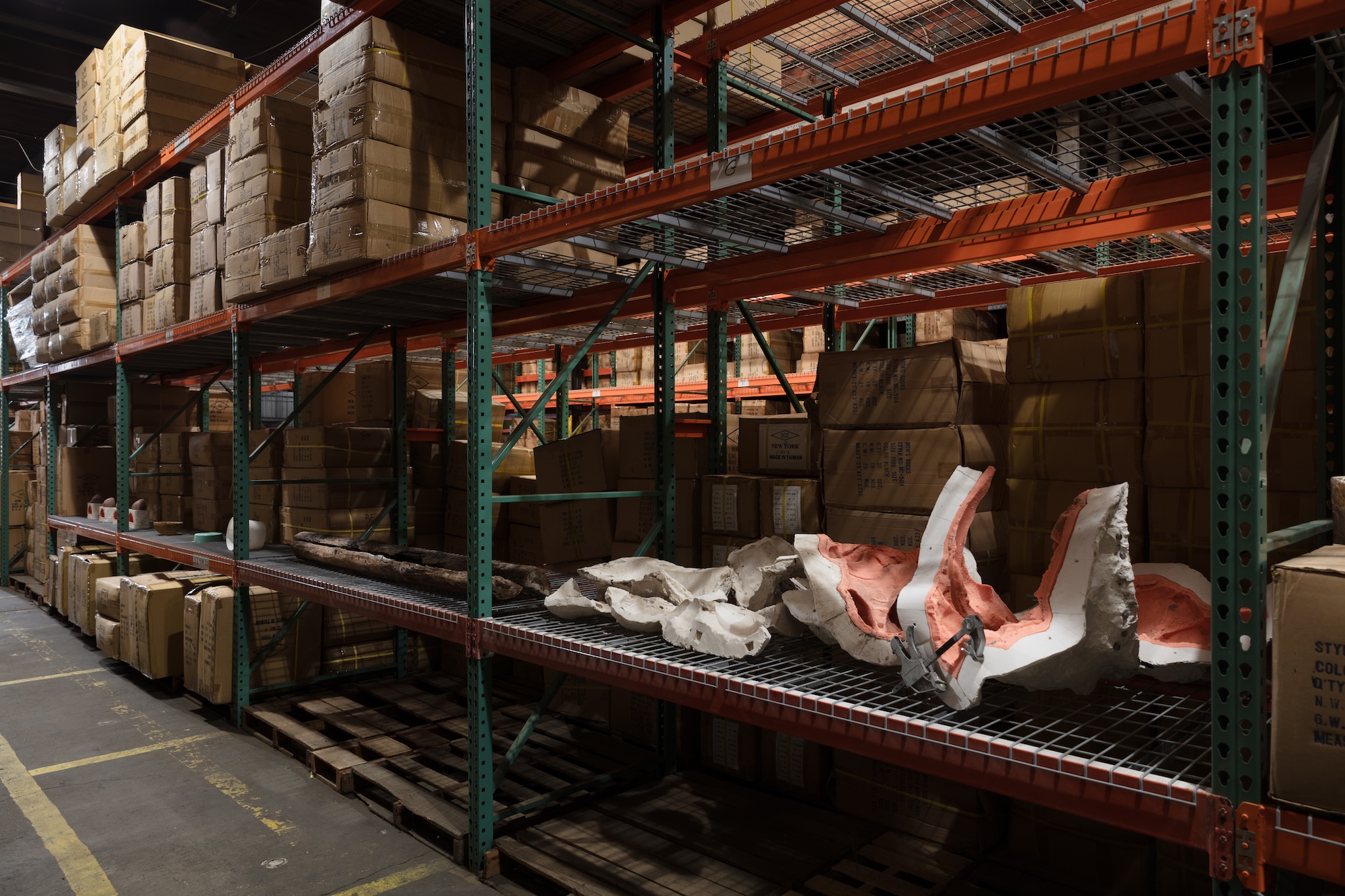A fragmented white and pink plaster or foam sculpture lies across a wire warehouse shelf amid stacked cardboard boxes.