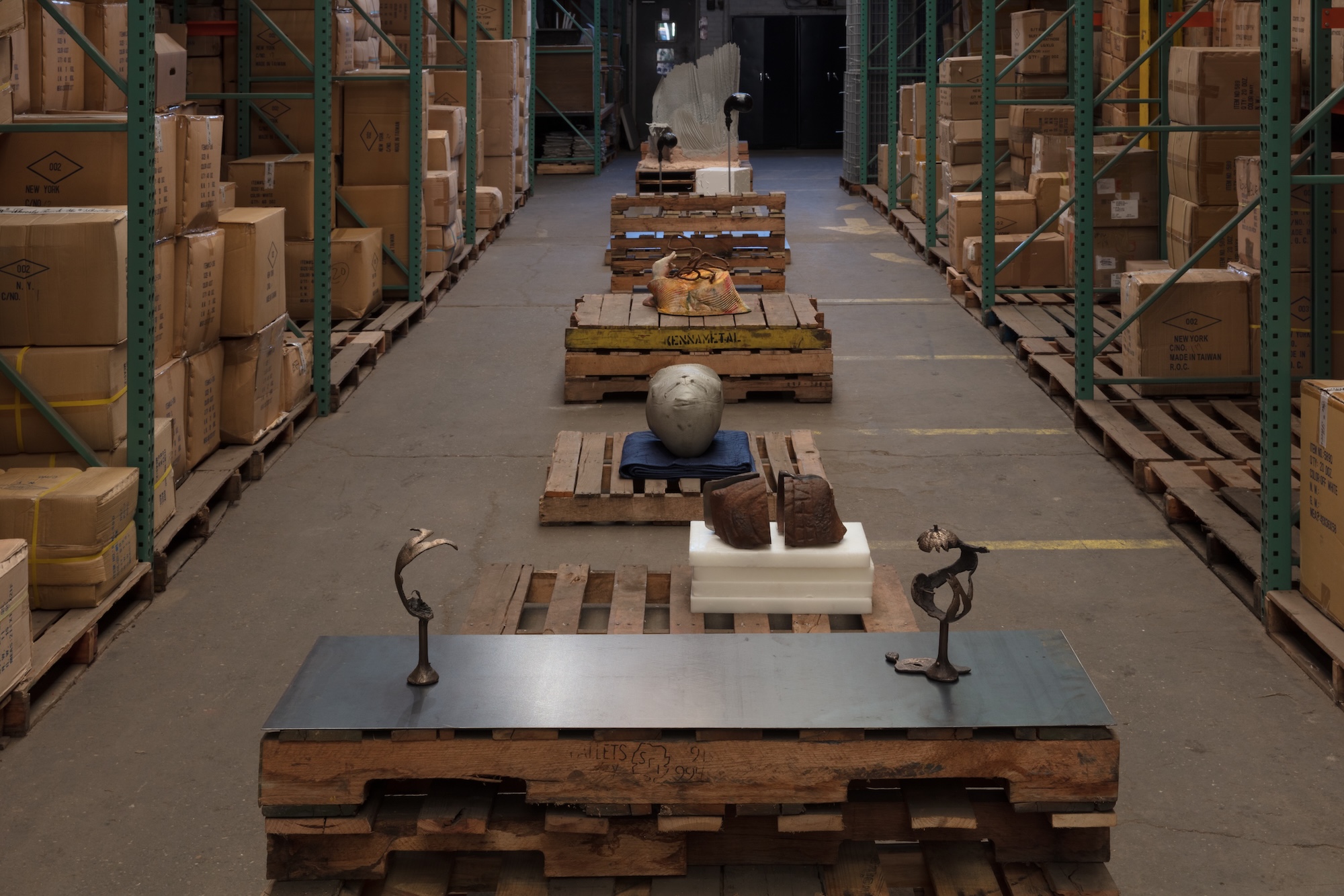 A warehouse aisle with sculptural objects on wooden pallets, including a steel plate with two small bronze figures, a sphere resting on folded fabric, and a large white wing-like form in the background.
