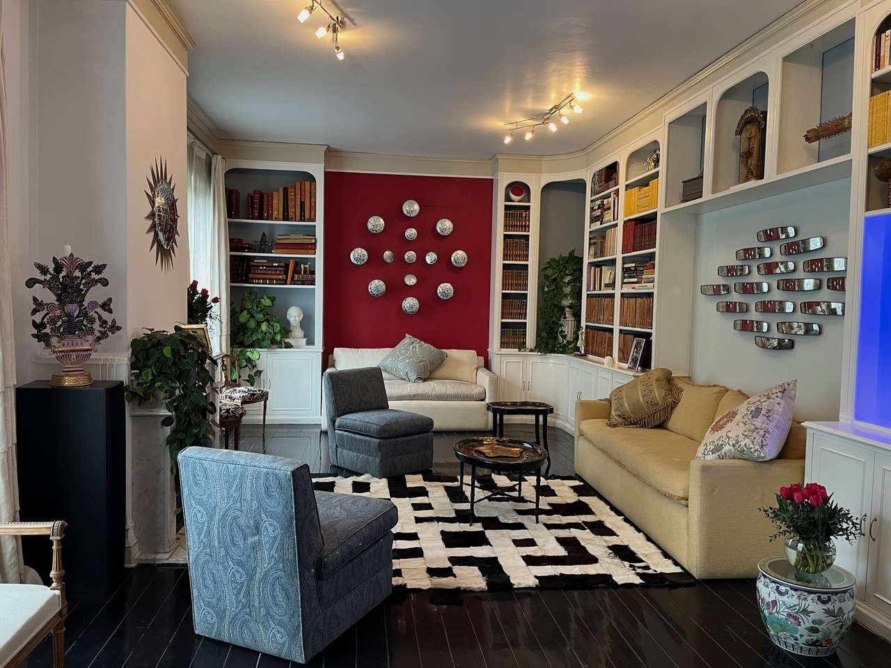 A sophisticated library-style living room with floor-to-ceiling bookshelves, a bold red accent wall adorned with silver decorative plates, eclectic seating, and a black-and-white cowhide rug.