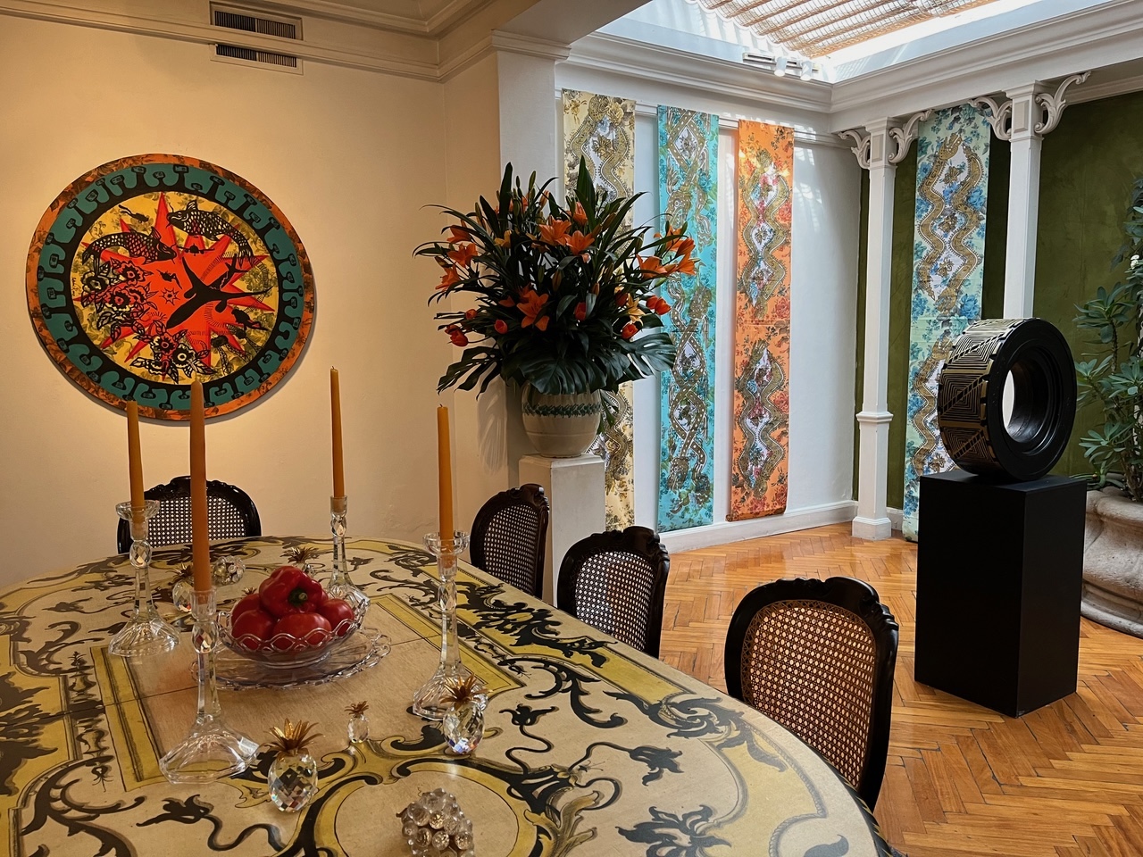 A elegantly decorated dining room featuring a vibrant circular artwork with fish motifs, colorful textile panels hanging between classical columns, and a ornate table set with candles and a bowl of red peppers.