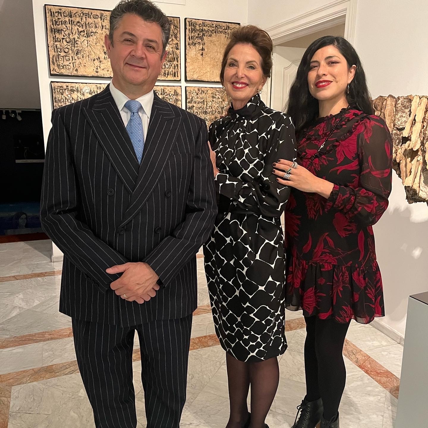 Three elegantly dressed people pose together at an art gallery opening, with large gold-leaf textual artworks displayed on the wall behind them.